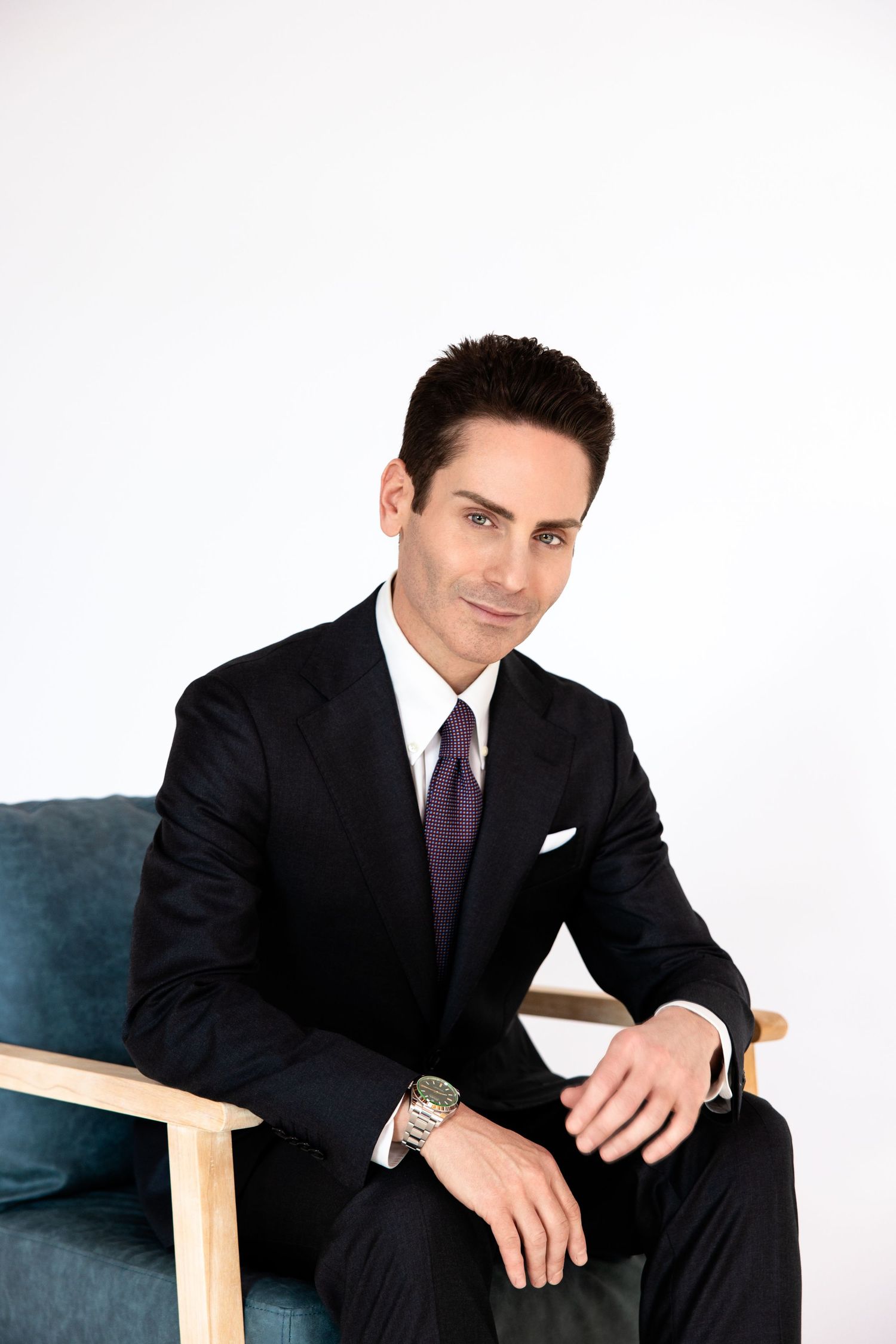 Professional man in black suit and purple tie seated in modern chair against white background.