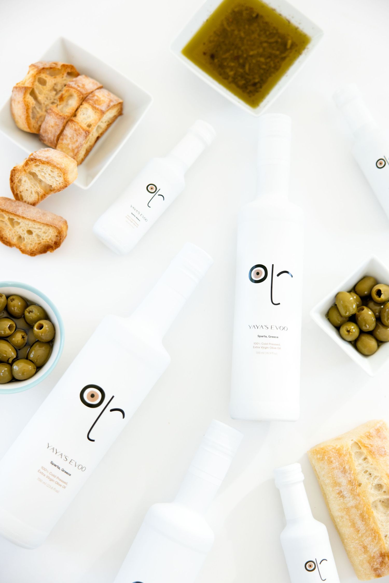 Overhead flat lay of bread slices, olives, and dipping oils in minimal white dishes on a bright background.