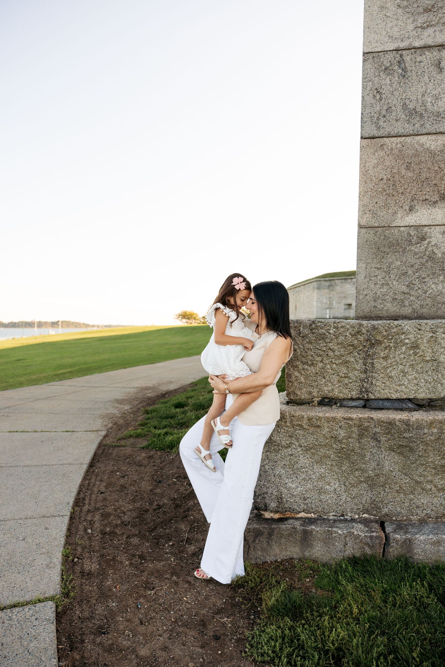 Mother and child share a tender moment at sunset by a stone wall on grassy grounds.