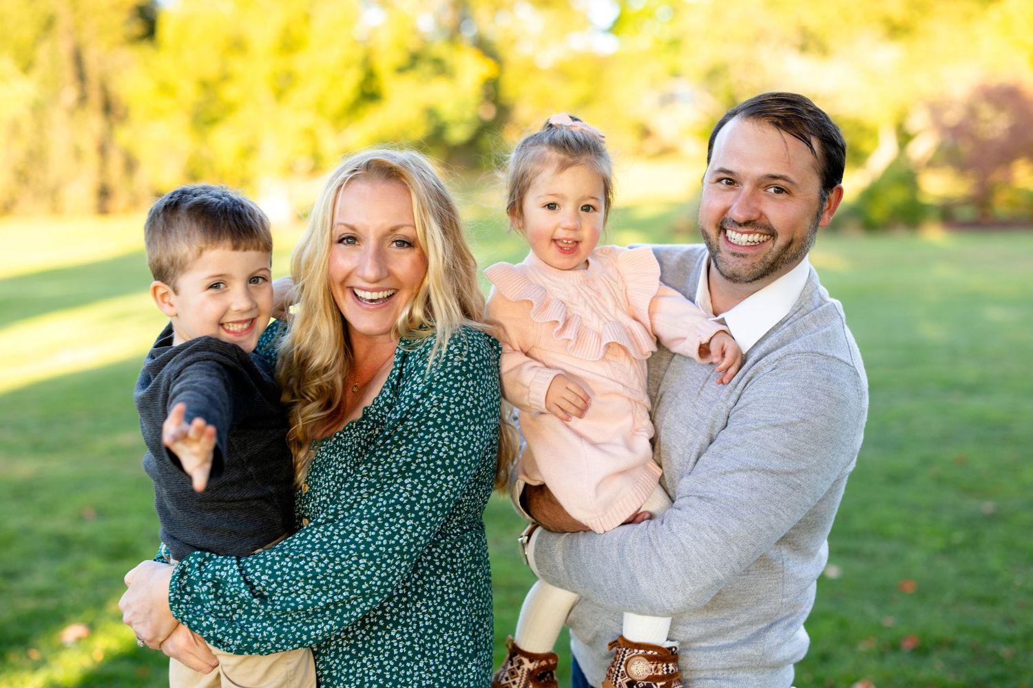 Family laughing and playing together during outdoor autumn photo session in a sunny park setting.