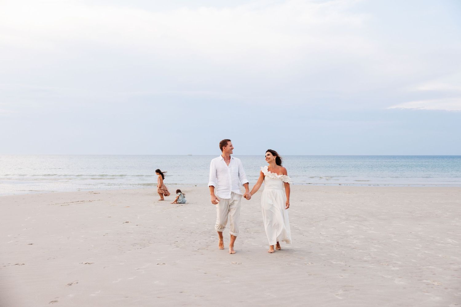 A couple walks hand in hand along a serene beach with calm ocean waters and cloudy skies in the background.