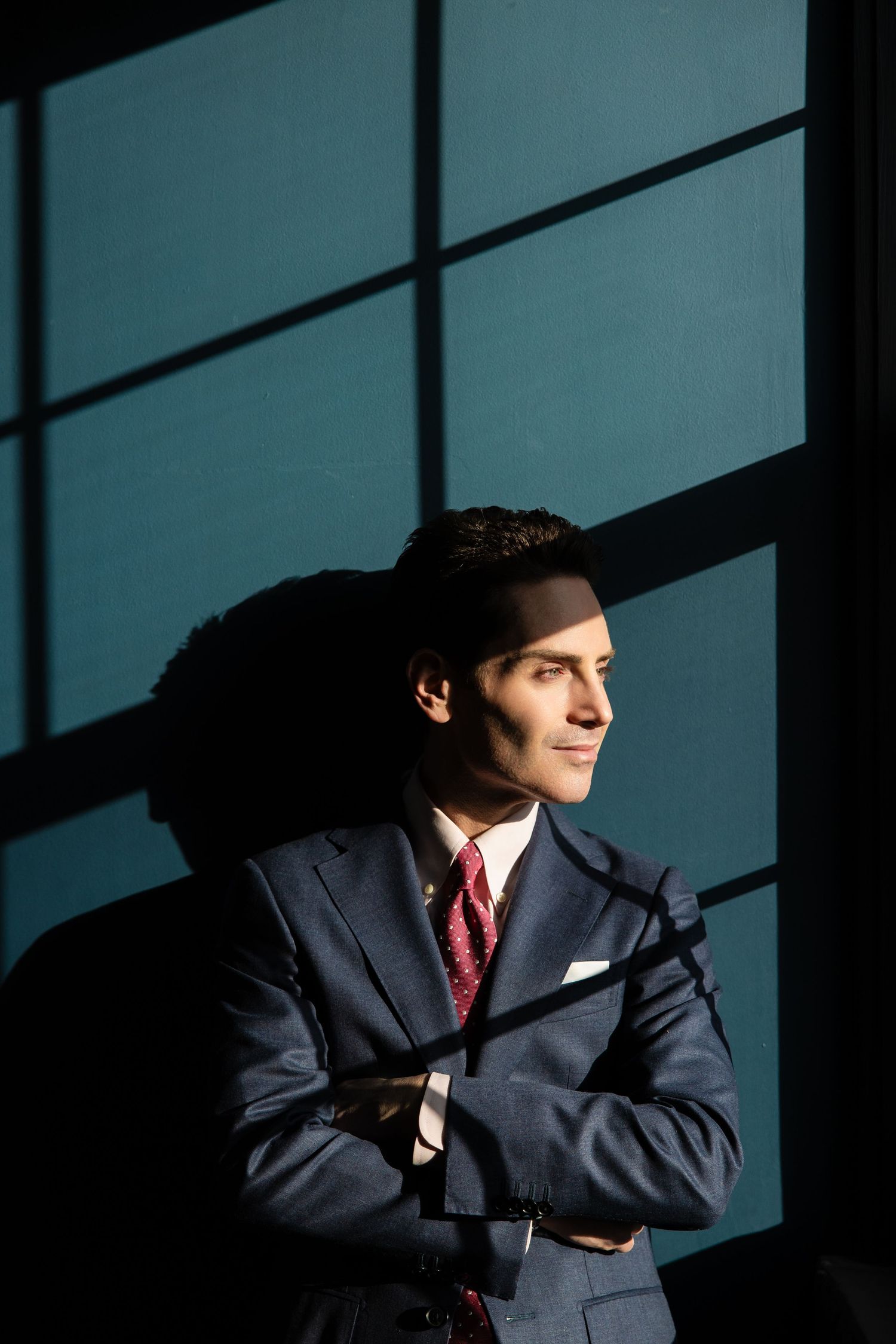 A confident young businessman in a navy suit and red tie stands with arms crossed, smiling in dramatic window light.