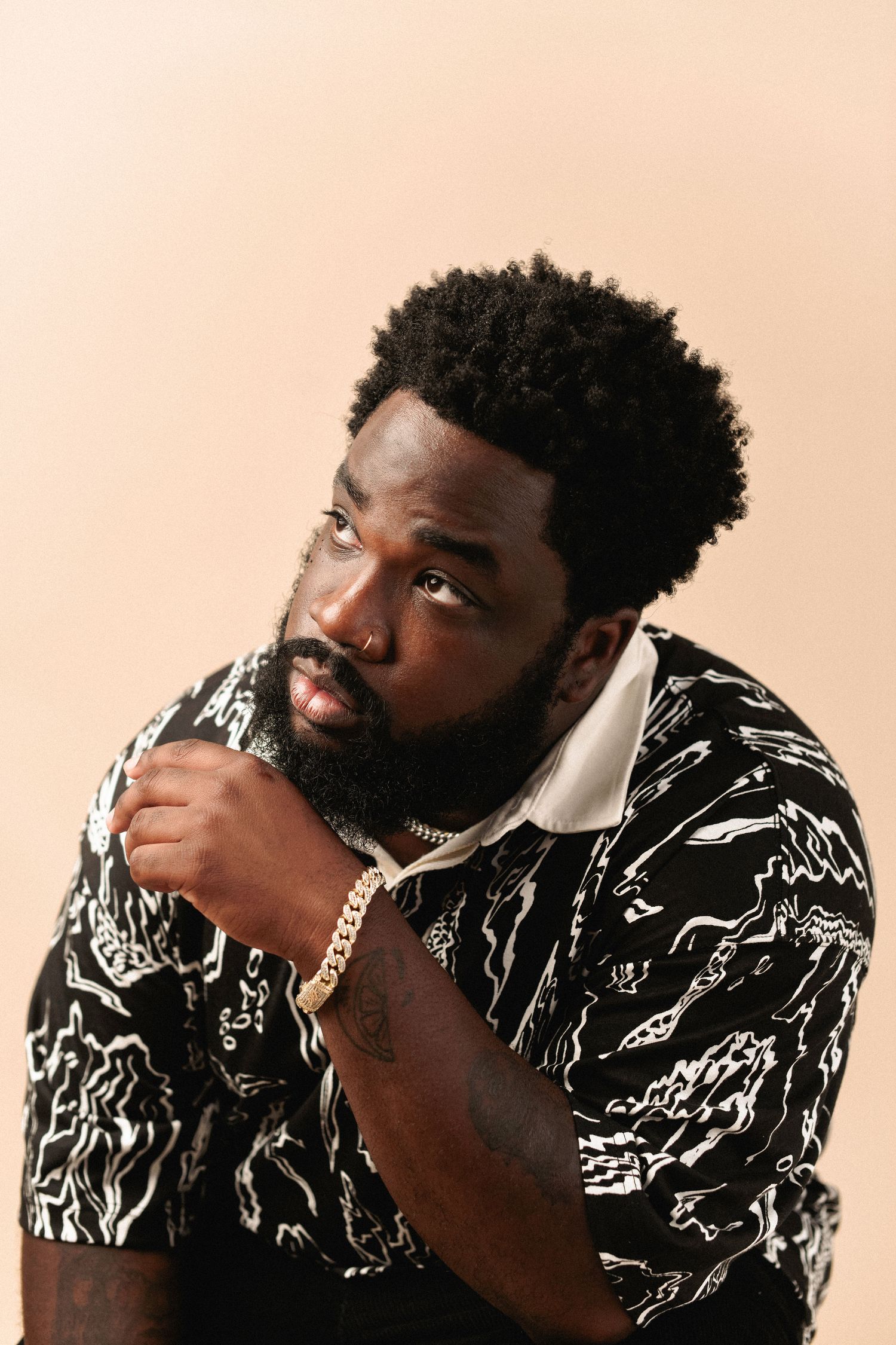 Confident bearded Black man in patterned shirt with gold chain posing against beige background.