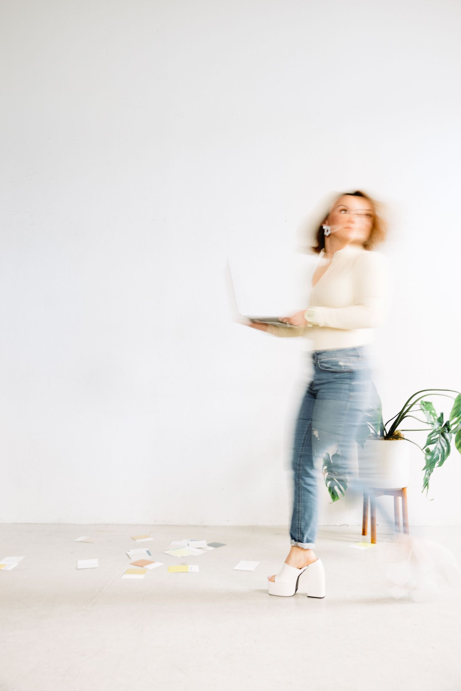 Blurred motion shot of a woman in jeans holding a laptop, standing near a potted plant on a white background.