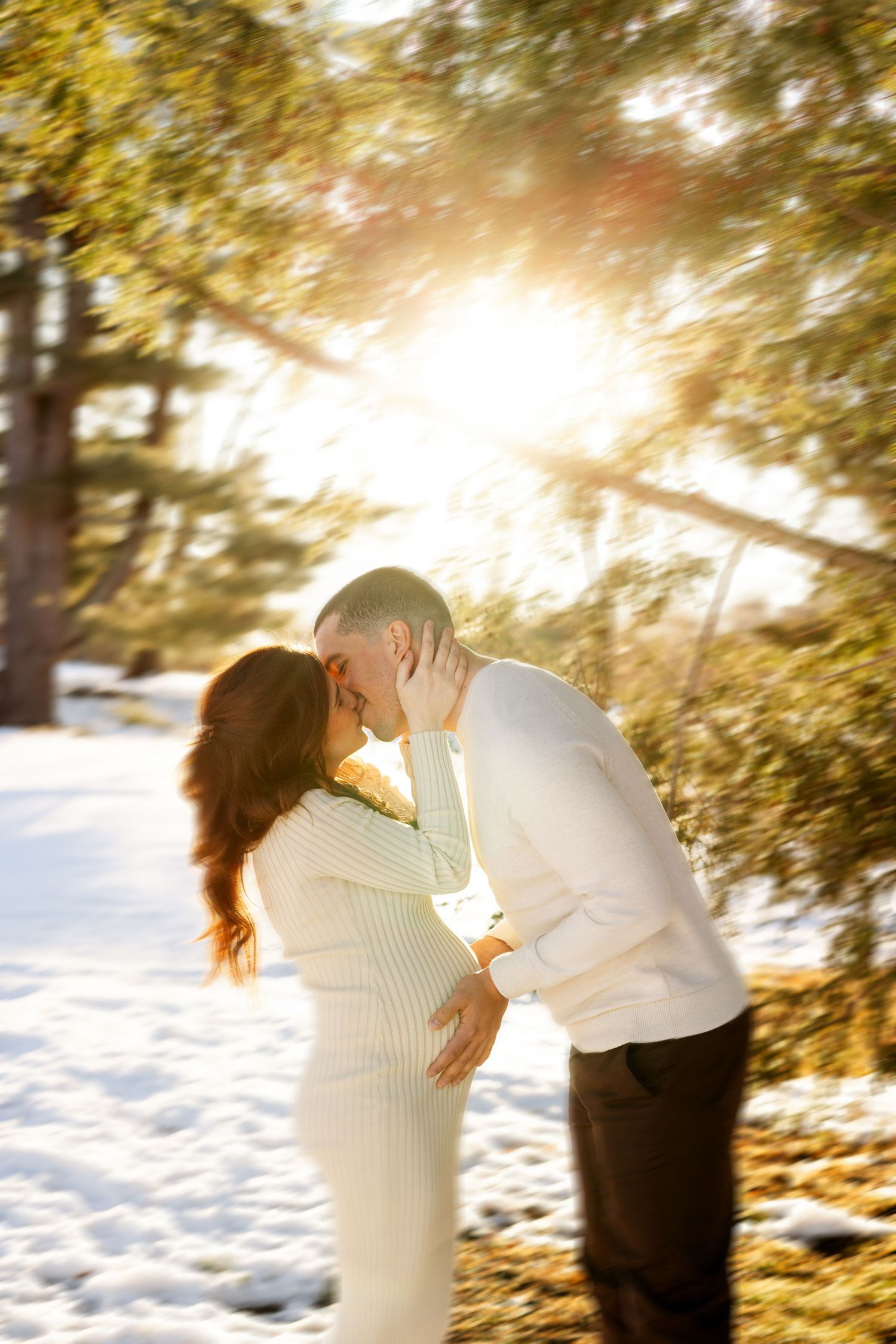 Pregnant couple kissing outdoors in snowy winter setting with golden sunlight filtering through evergreen trees.