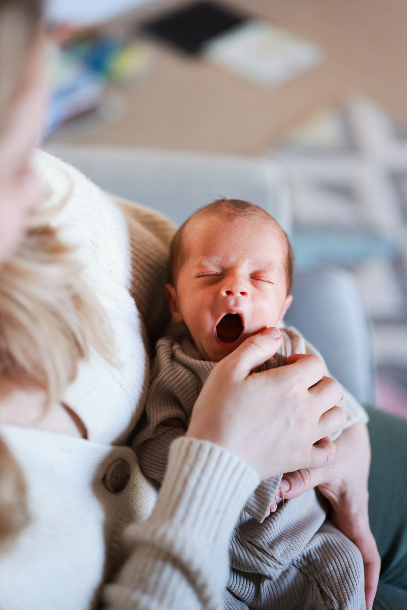 Yawning newborn baby swaddled in white blanket lying on bed being held gently by caregiver wearing sweater.