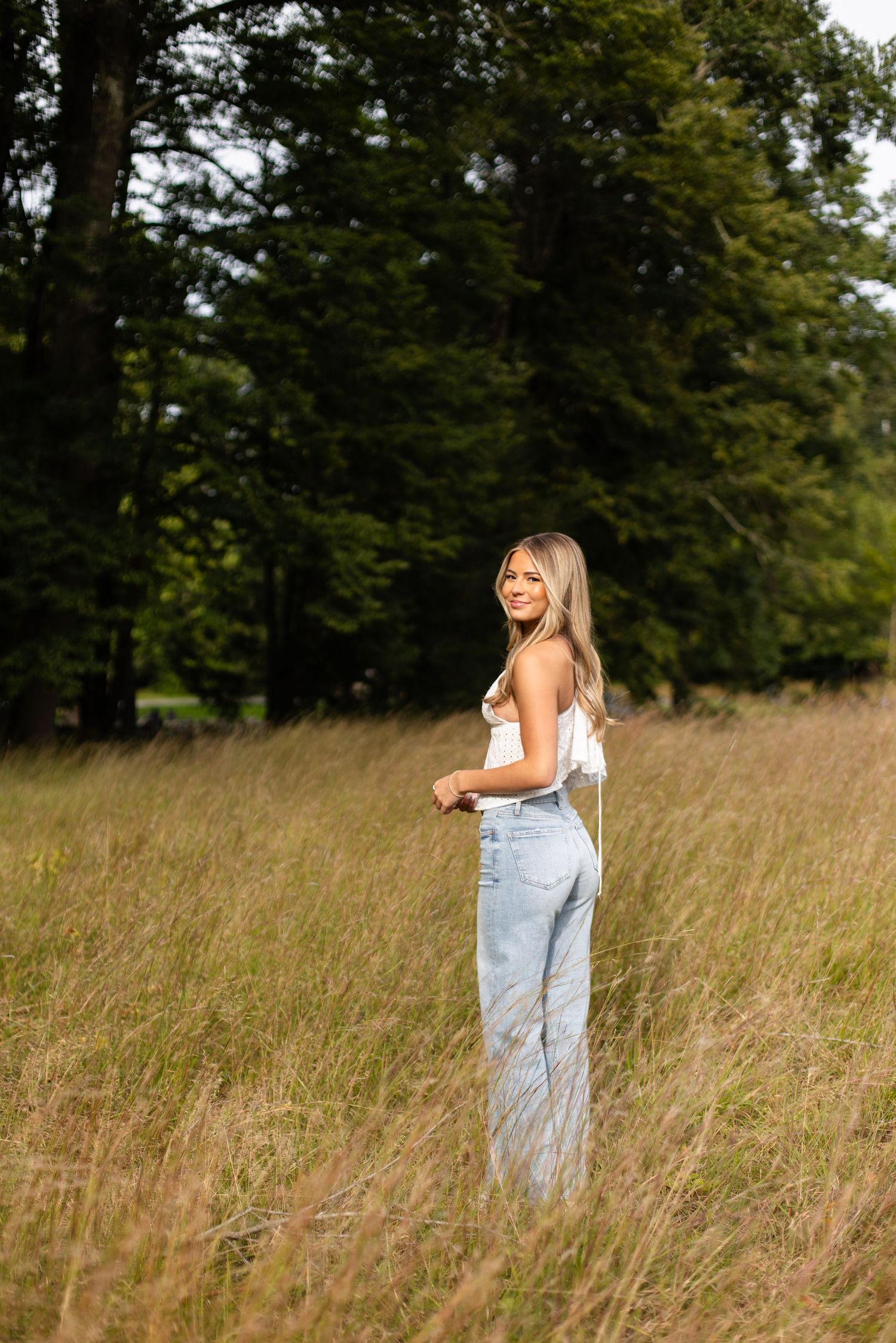 A person in light blue jeans and a white top stands in a sunlit meadow with tall grass and trees in the background.