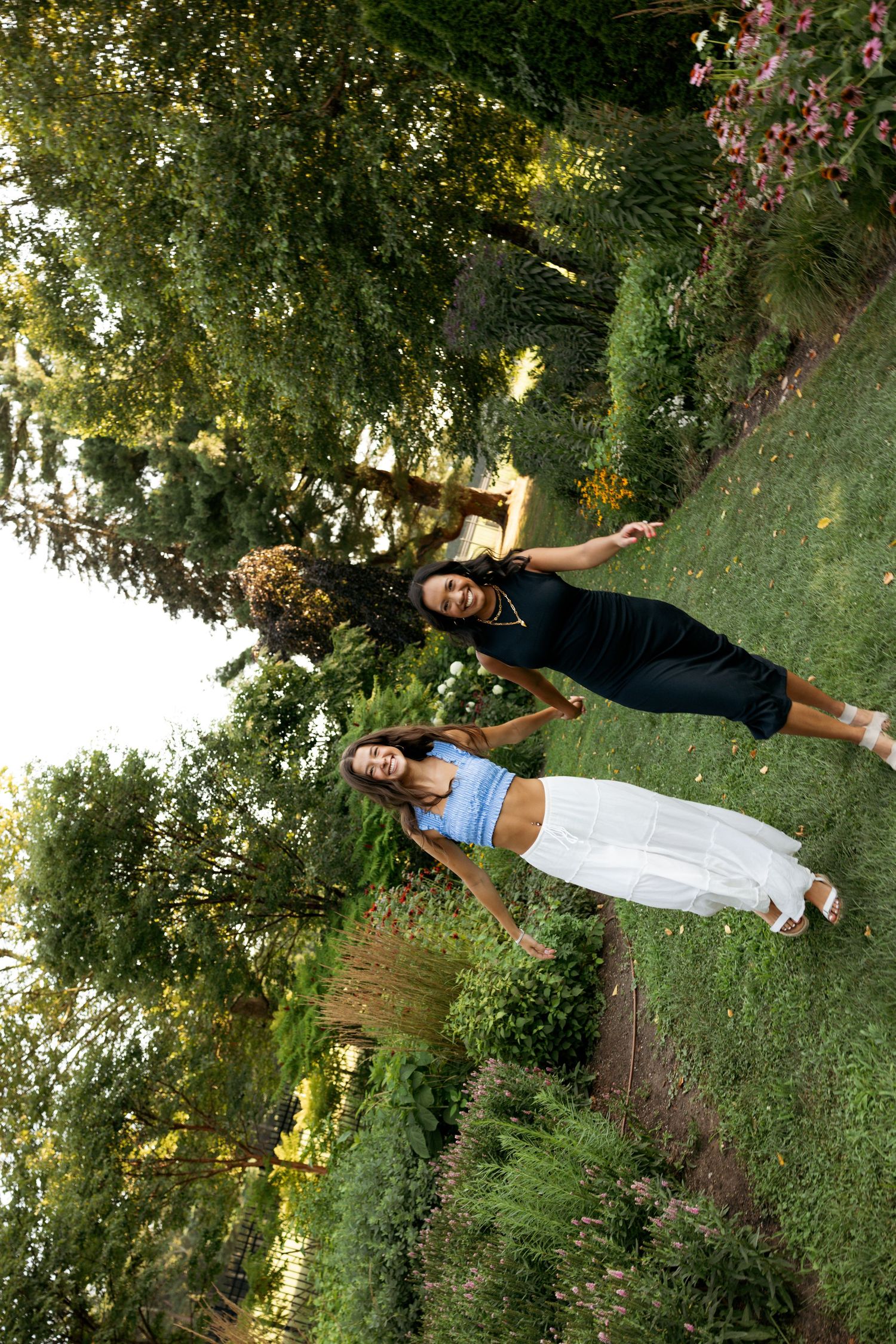 Three people strike joyful poses on a grassy lawn with trees and flowers in the background.