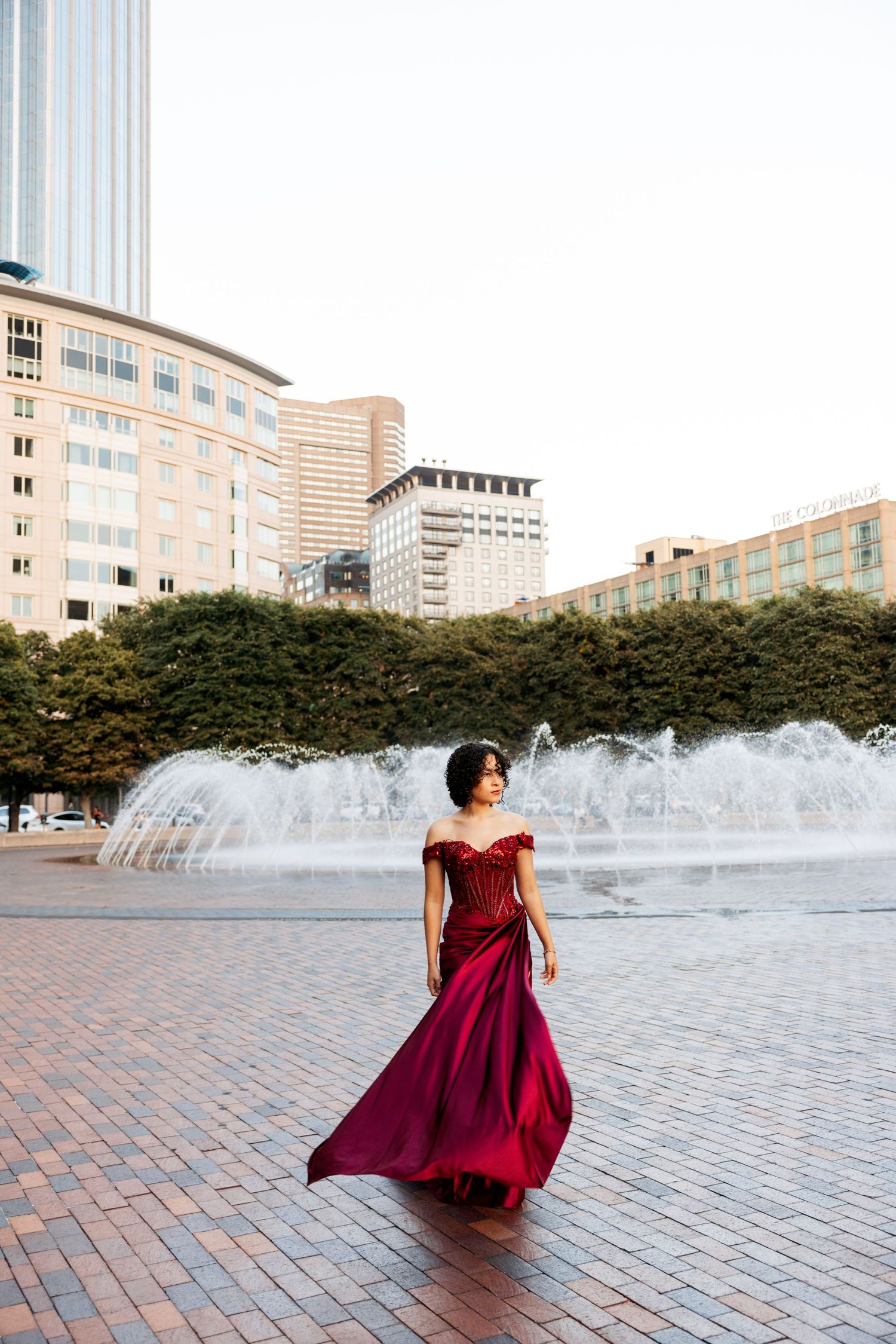 A flowing burgundy evening gown twirls gracefully against an urban backdrop with fountains and modern buildings.