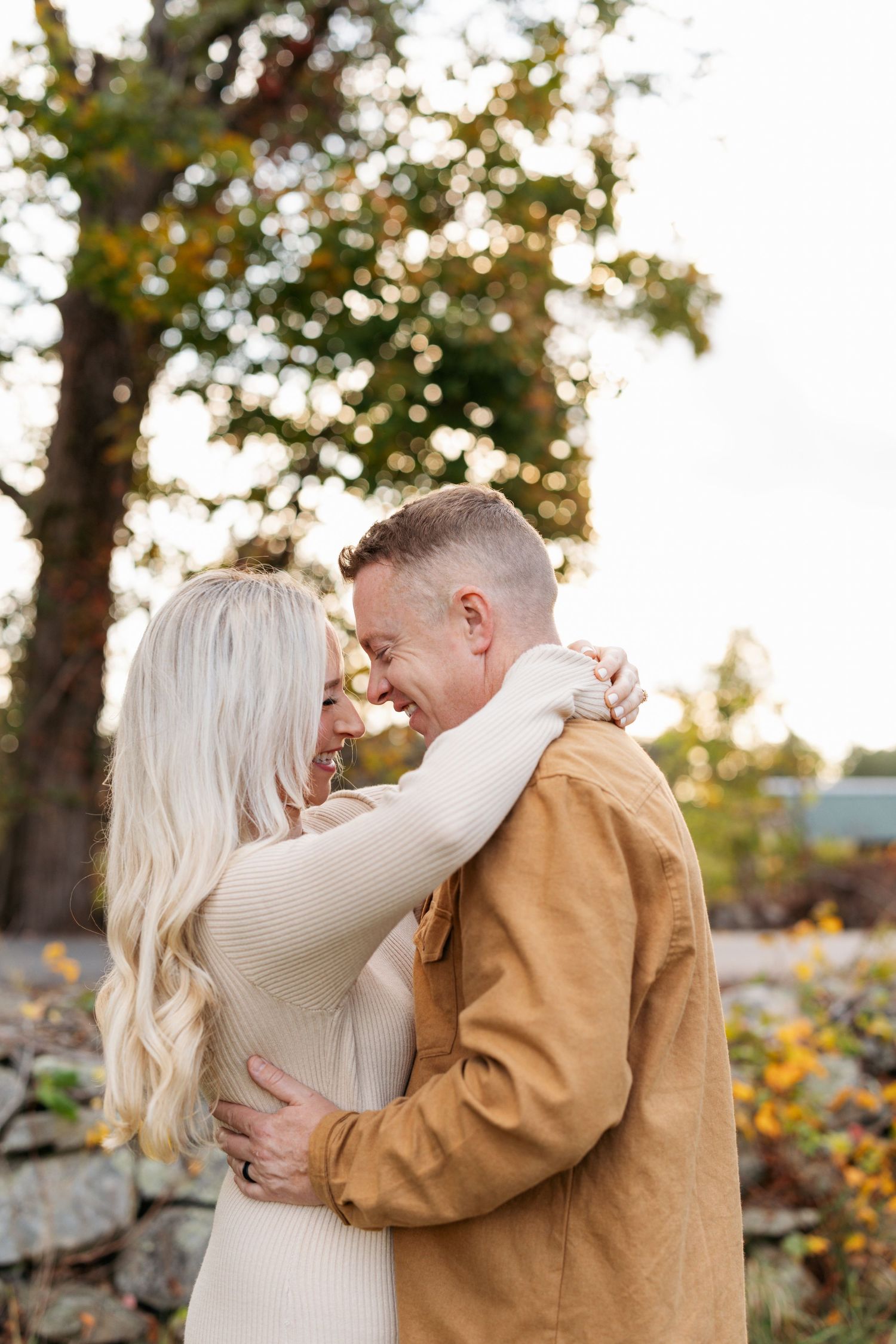 A couple shares a romantic embrace outdoors surrounded by autumn trees and golden sunlight.