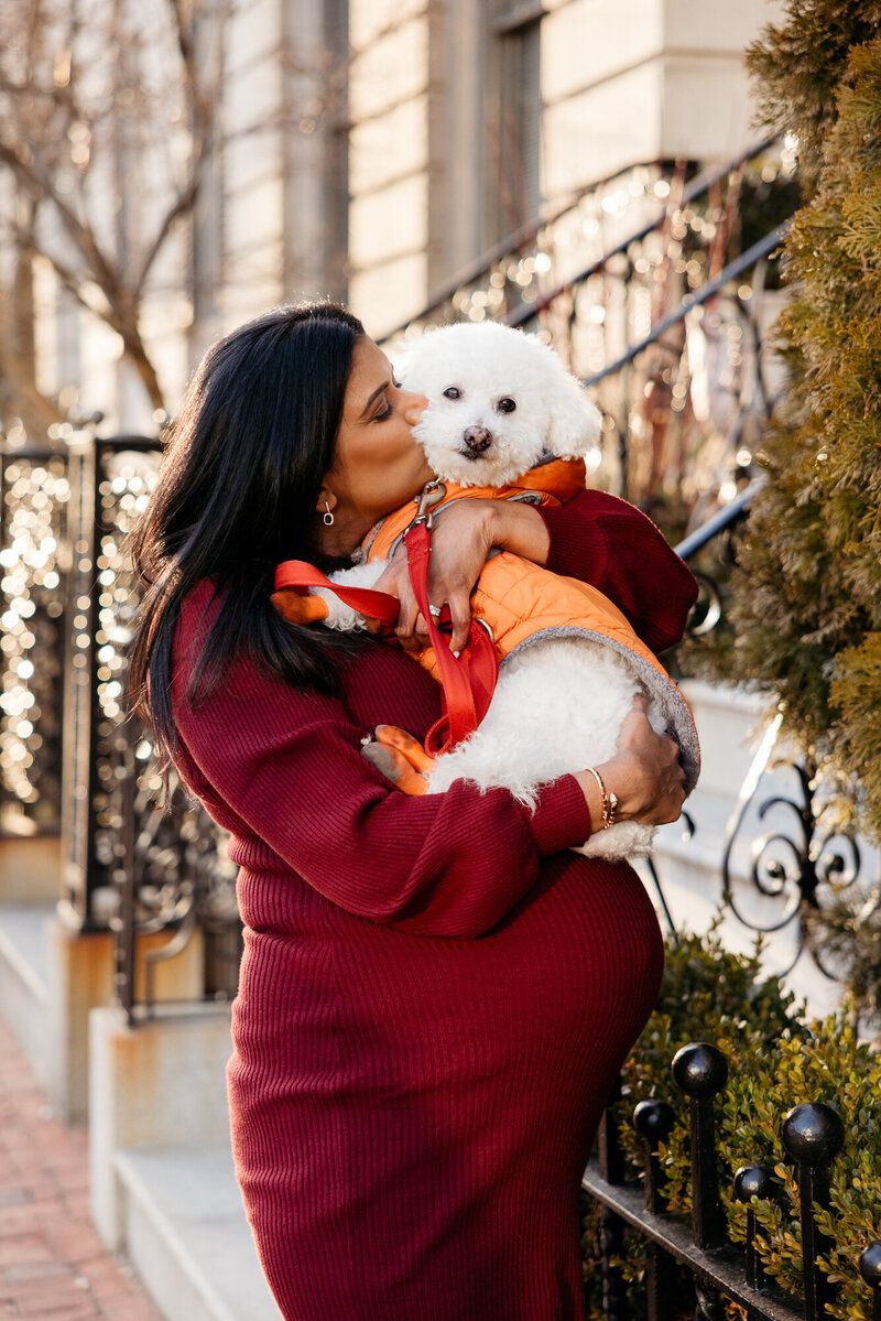 Person in burgundy dress holding and kissing a small white fluffy dog on urban steps with holiday decorations.