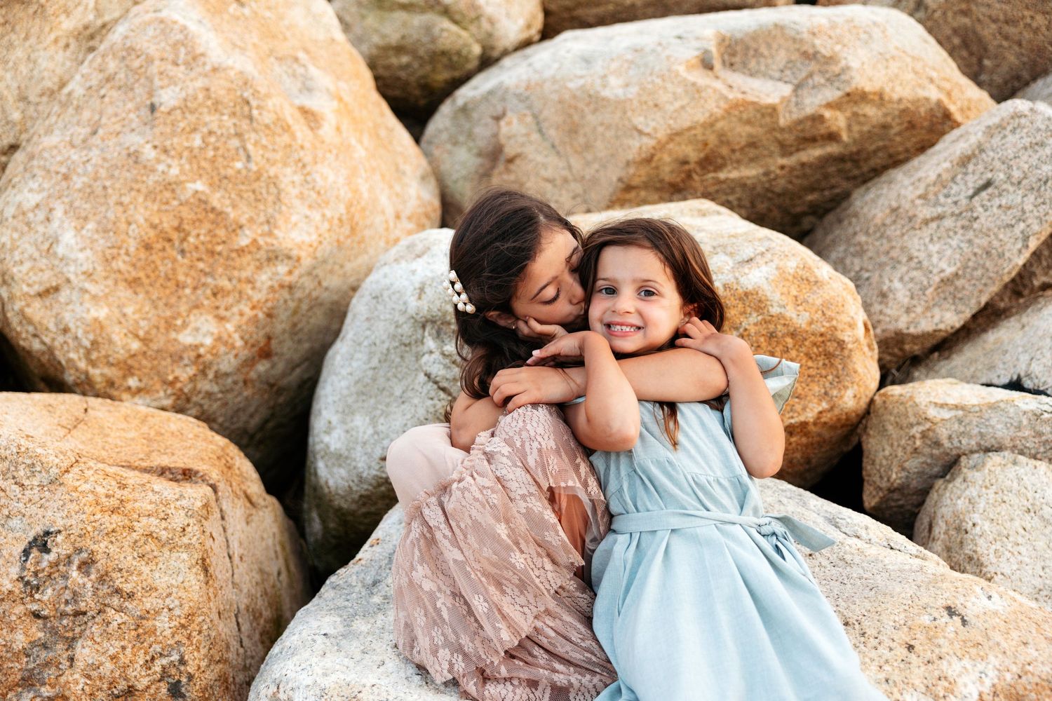 Family group poses intimately among large coastal rocks wearing coordinated pastel outfits for a beach photo session.