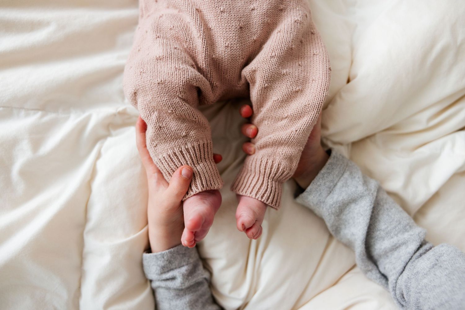 A baby wearing a pink sweater grasps a parent's finger while lying on white bedding.
