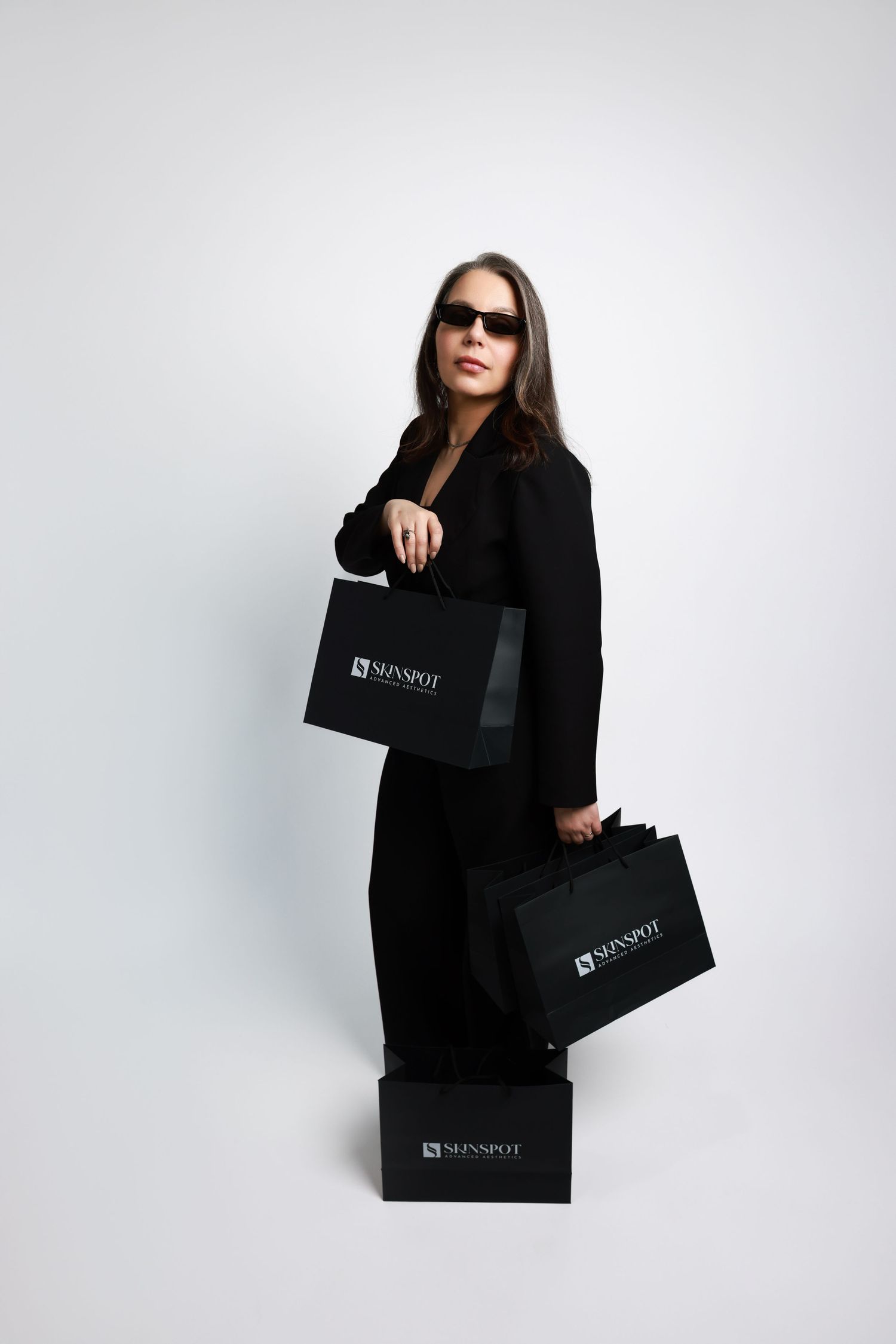 Stylish figure in black outfit poses with multiple shopping bags against white backdrop.