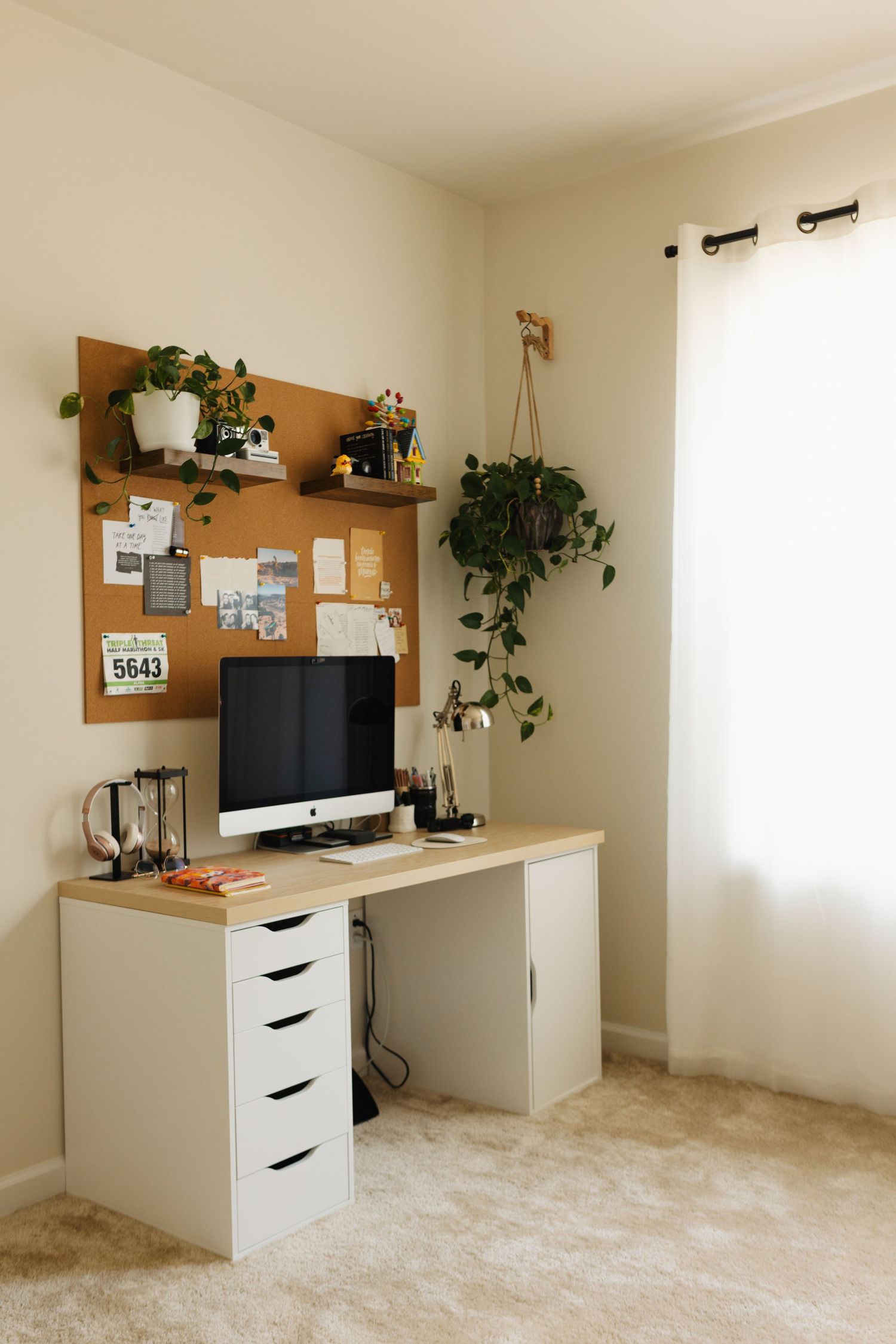 Modern home office setup with white desk, iMac computer, cork board, and hanging plants against beige wall and carpet.