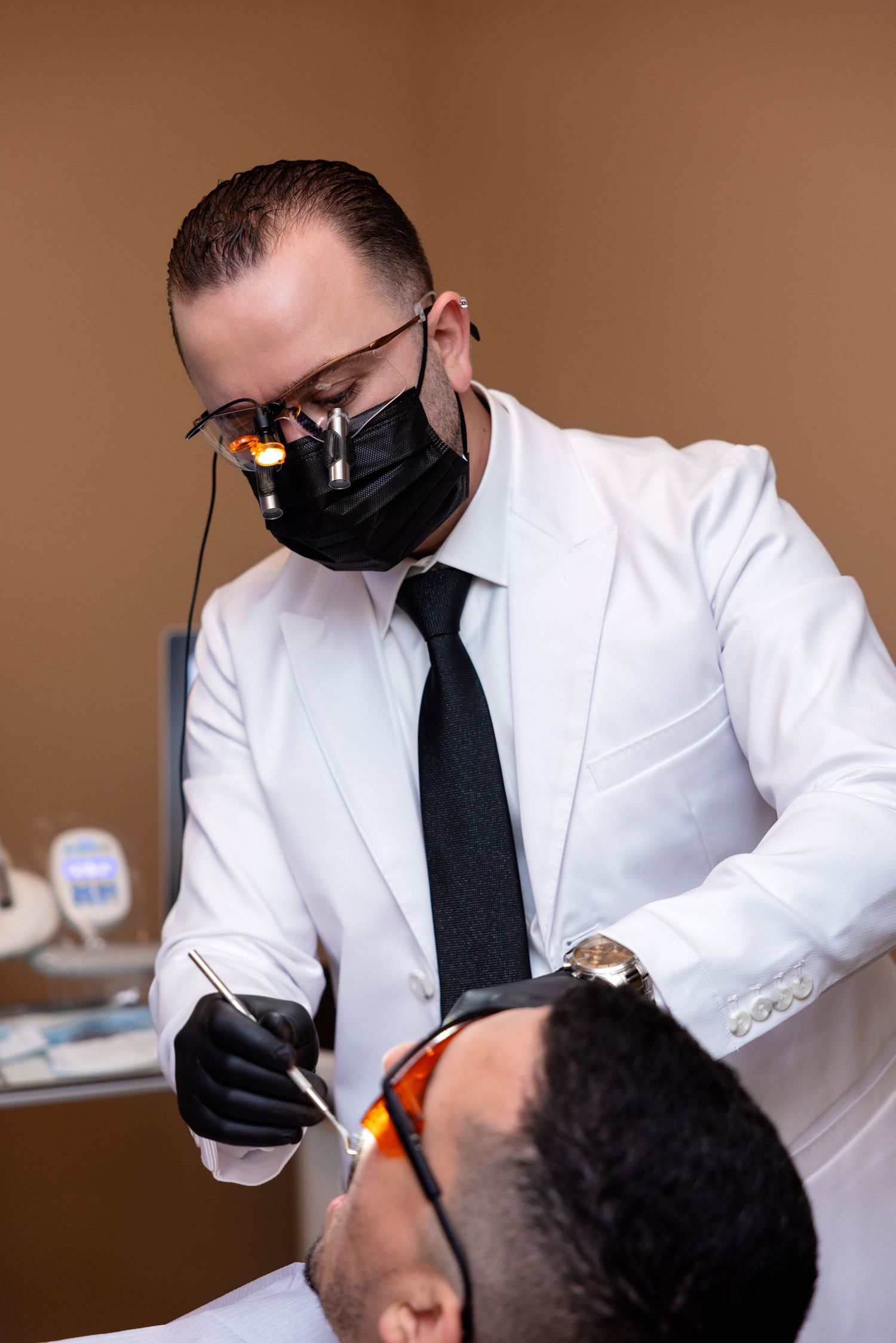Dentist in white coat and black mask uses dental tools on a patient wearing protective orange glasses in a clinic.