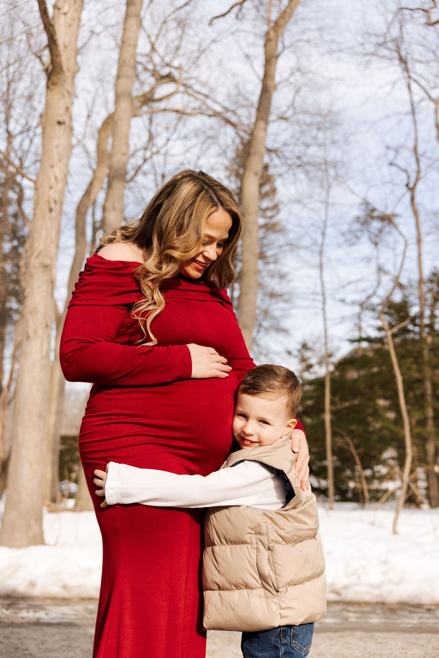 Winter maternity photos featuring red dress against snowy woodland backdrop.