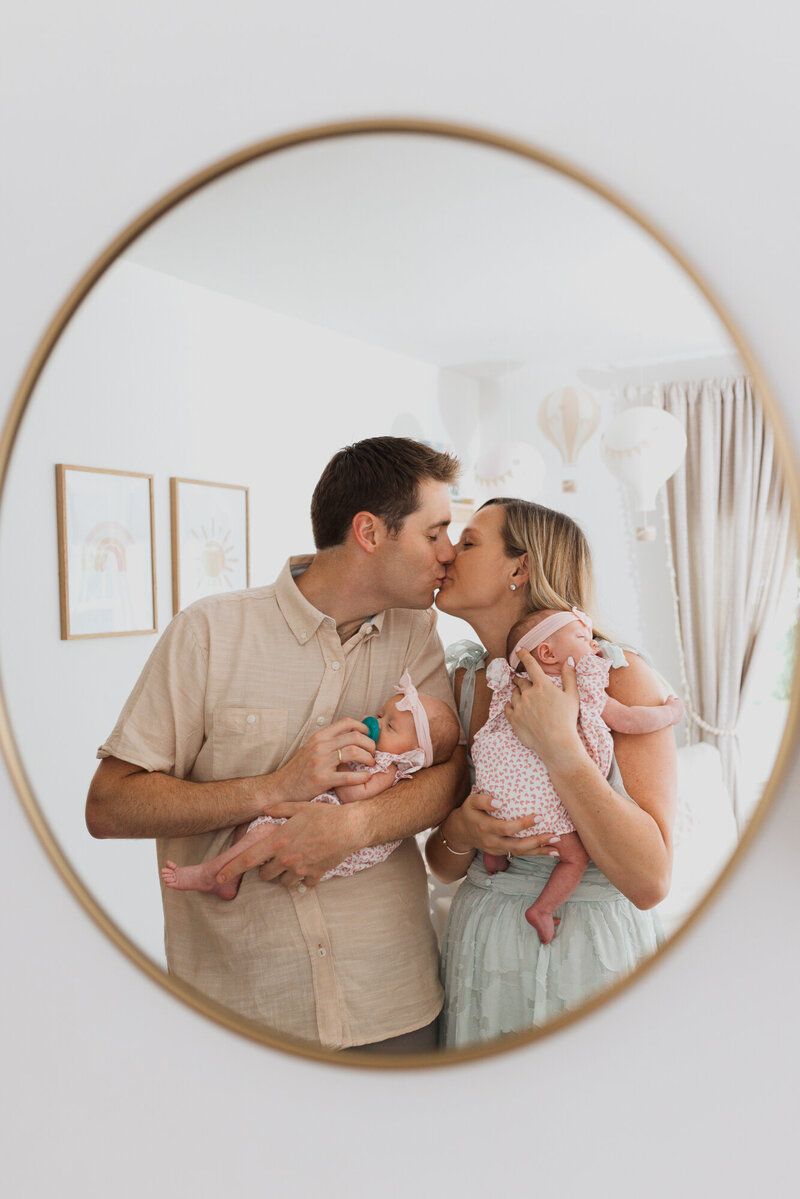 A tender family moment captured in a round mirror reflection showing parents holding and kissing their baby.