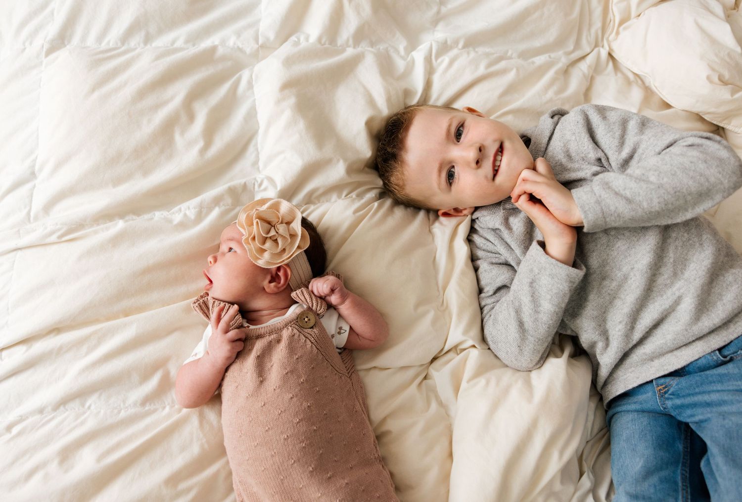 A baby lying on their back wearing a gray sweater smiles and giggles playfully on a white bed.