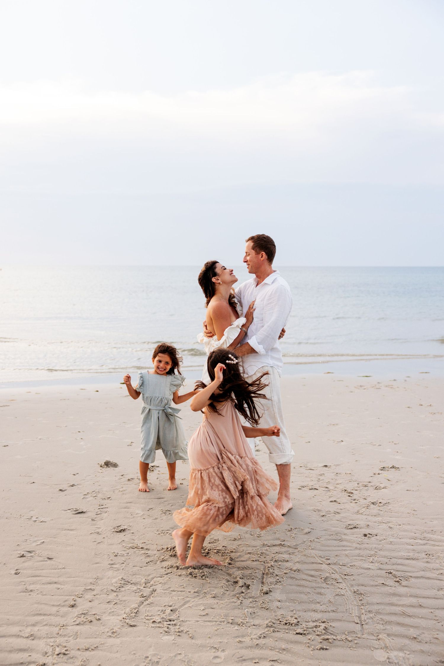 Family in pastel dresses and white shirt share joyful moments while dancing together on the beach.