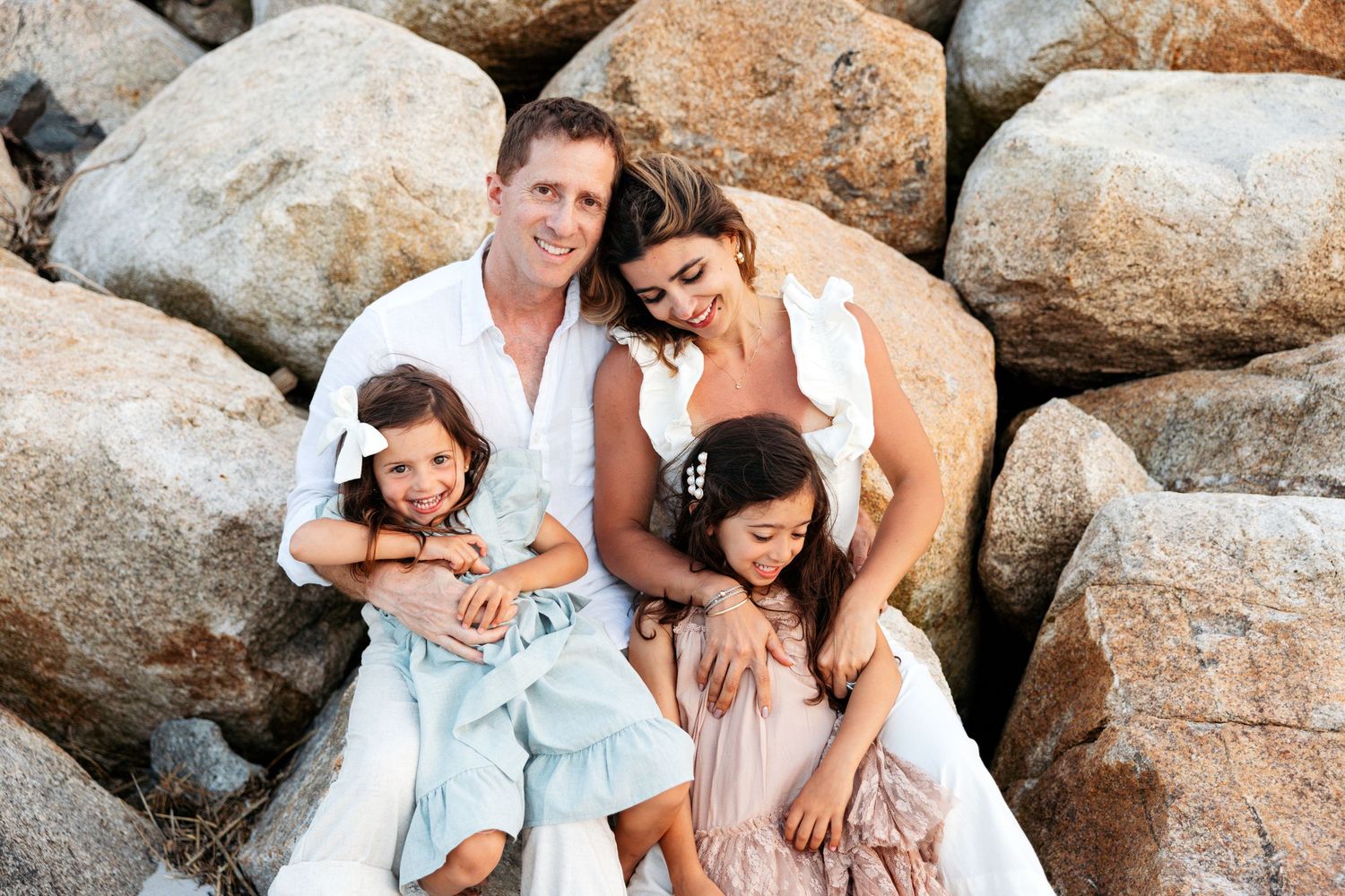 A family dressed in light colors sits together on coastal rocks while having their portrait taken at sunset.