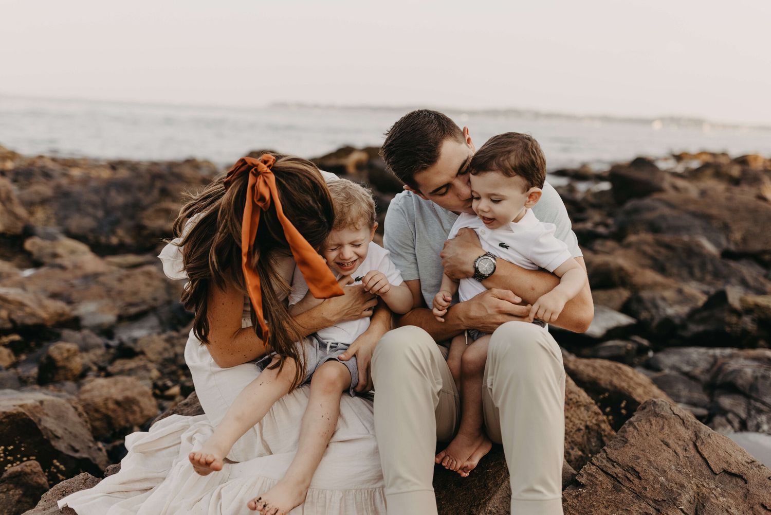Family sitting together on rocky shoreline during sunset photoshoot.