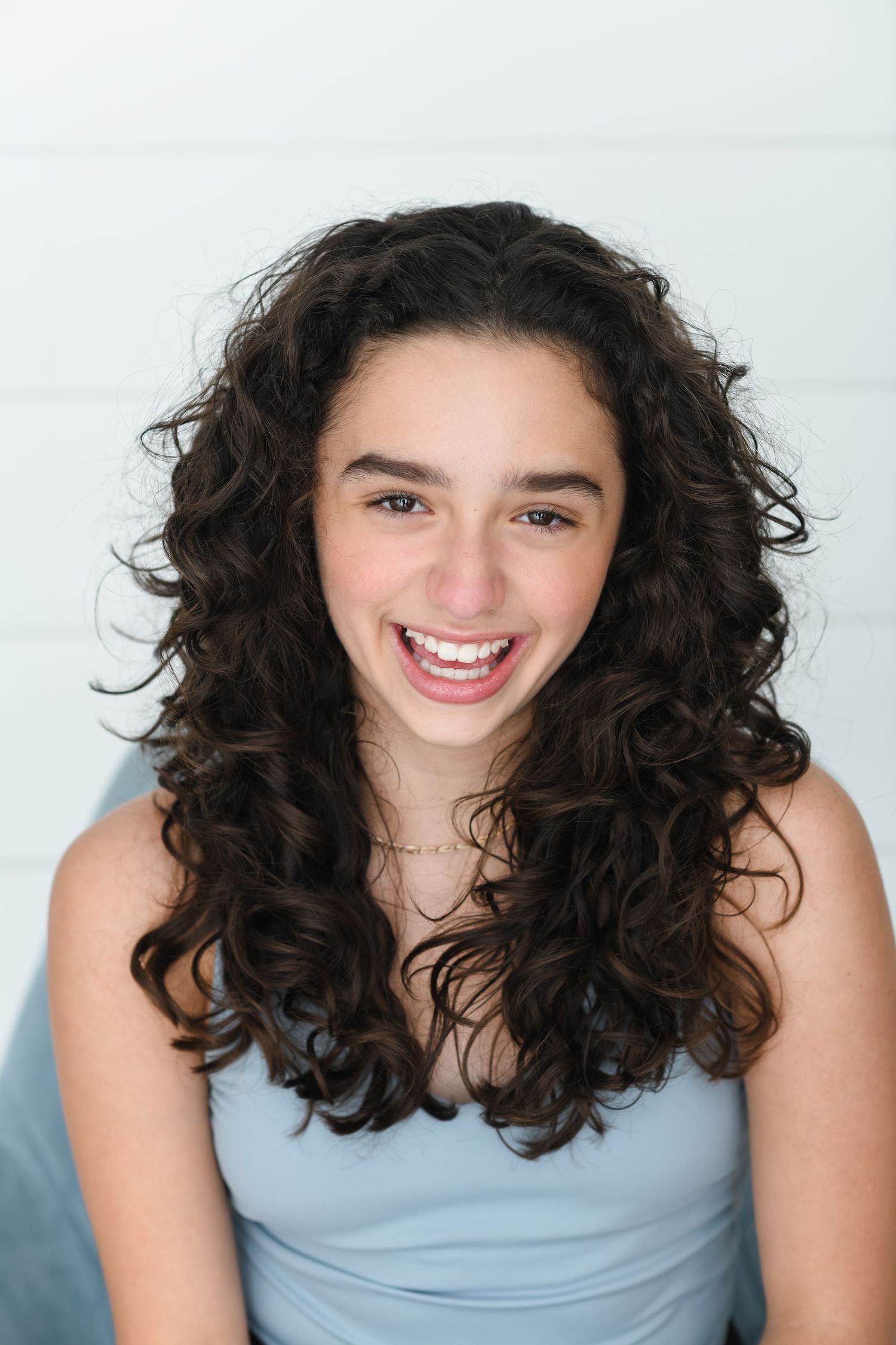 A teenage girl with long curly brown hair smiles brightly in a light blue top against a white background.
