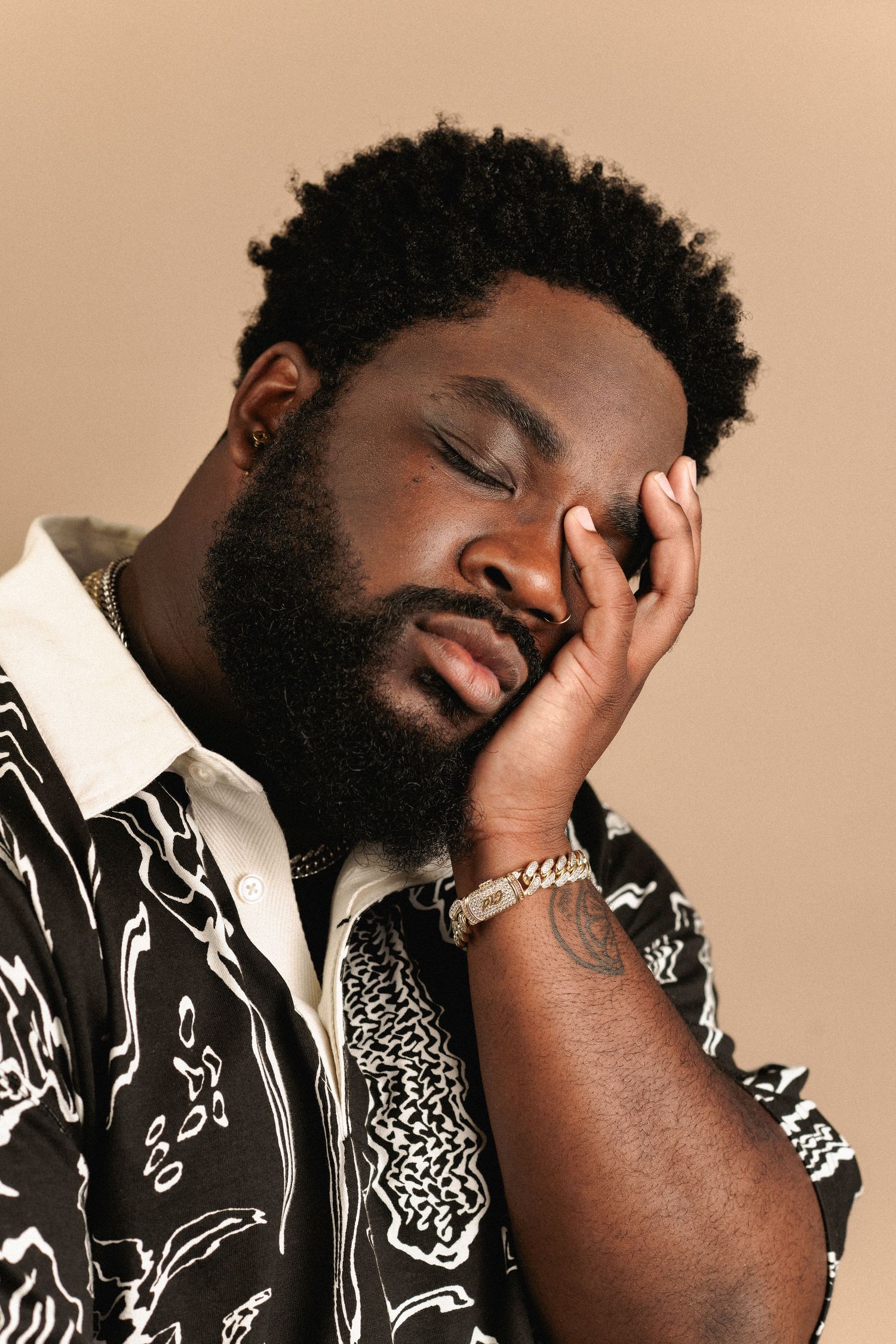 Bearded Black man in patterned shirt resting face in hand with eyes closed on beige background.