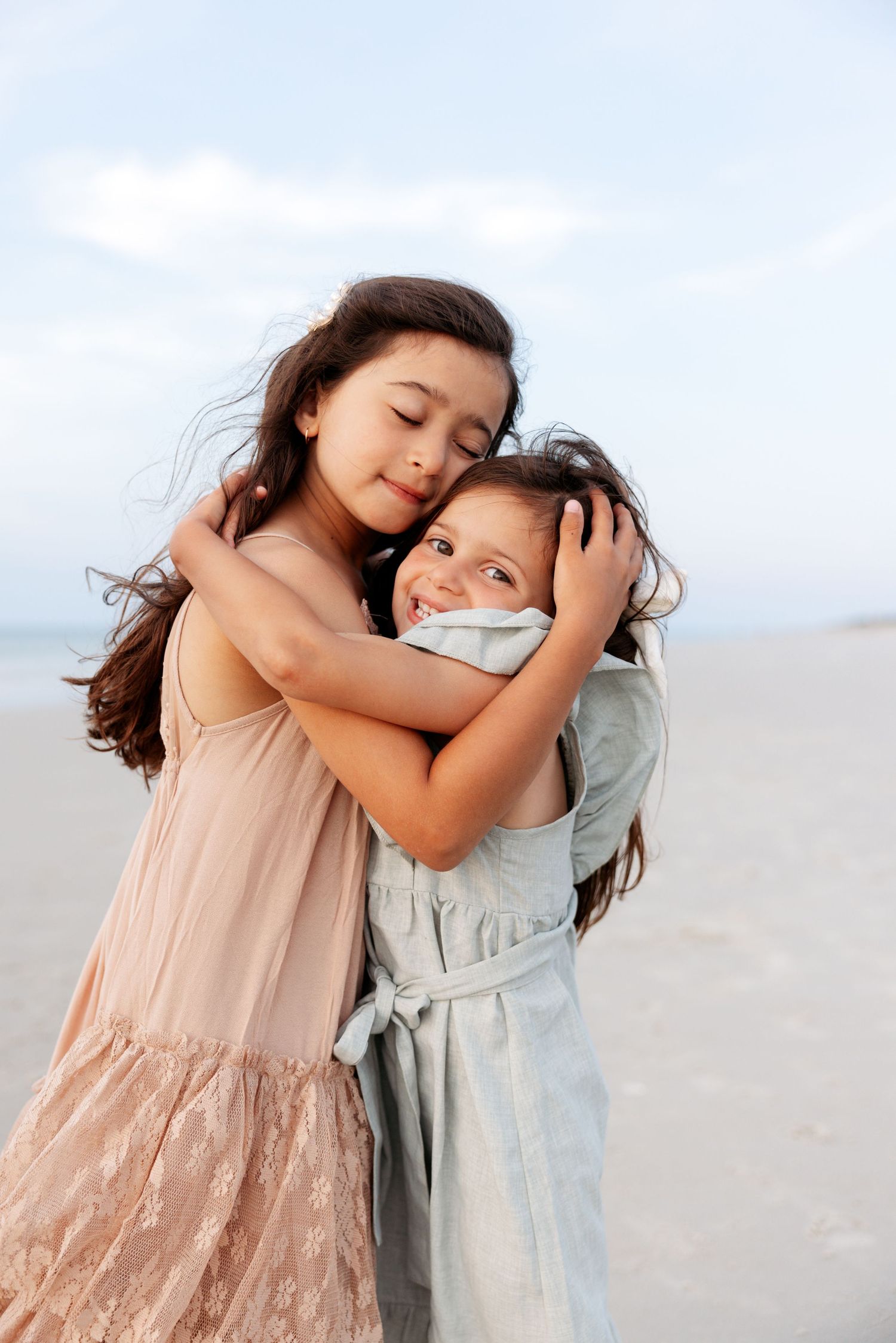 Children embrace and share affectionate moments while wearing pastel dresses against a light sky background.
