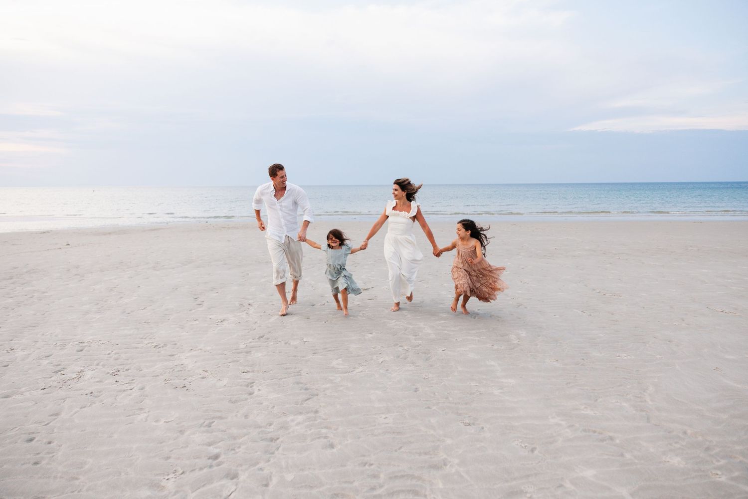 Family walking and playing together on white sandy beach with blue ocean waters in background.
