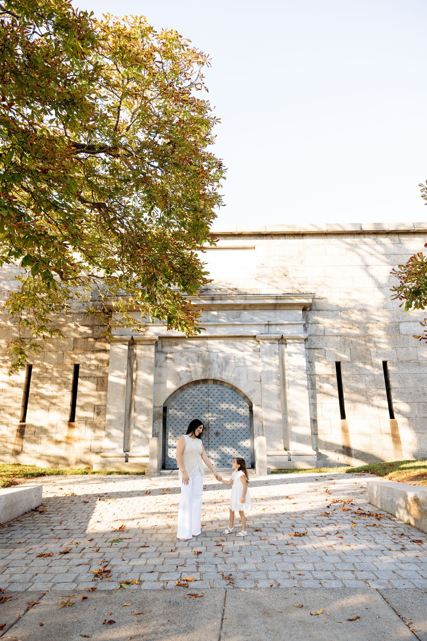 Stone wall with arched doorways casts dramatic shadows on cobblestone path while autumn leaves fall from tree above.