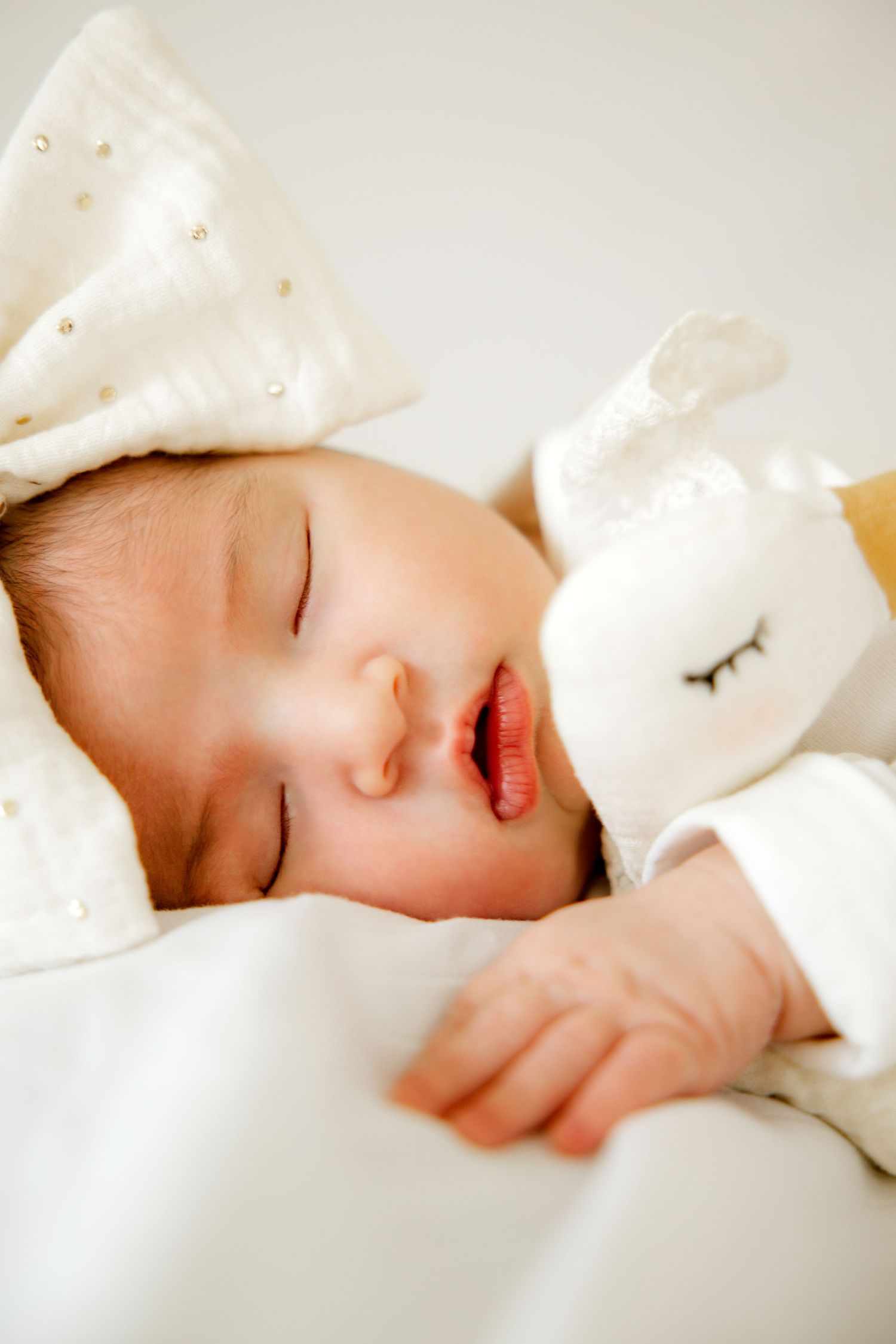 Peaceful newborn baby sleeping with a white stuffed bunny toy, wearing a white outfit with a bow headband.
