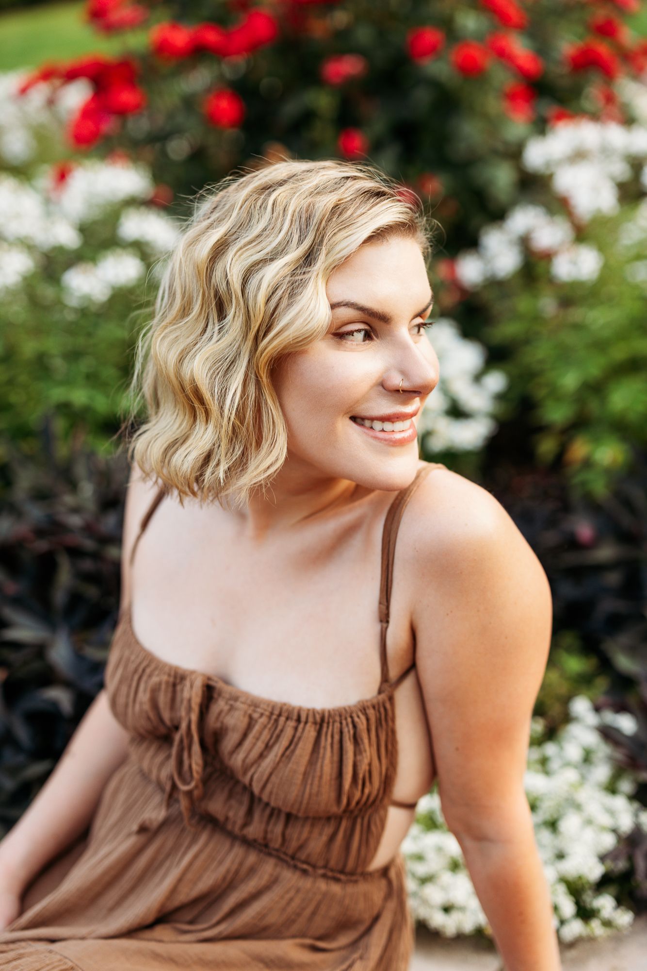 A woman in a brown spaghetti strap dress poses outdoors among blooming red and white flowers in summer.