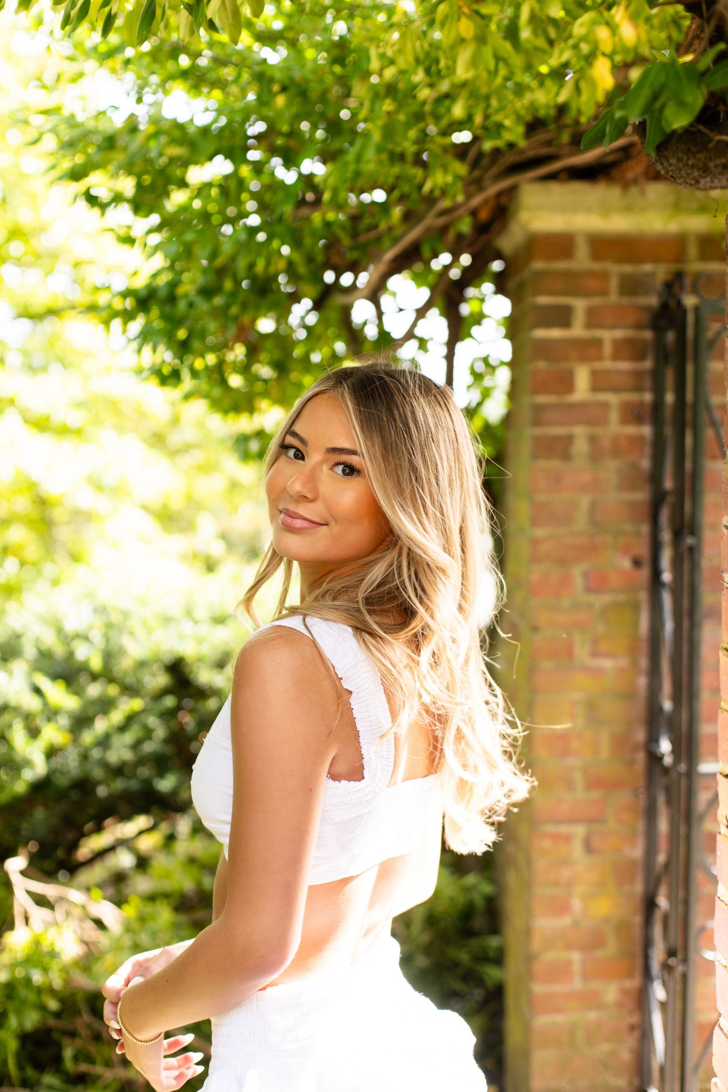A woman in a white dress poses elegantly against a brick wall backdrop with sunlit foliage in the background.