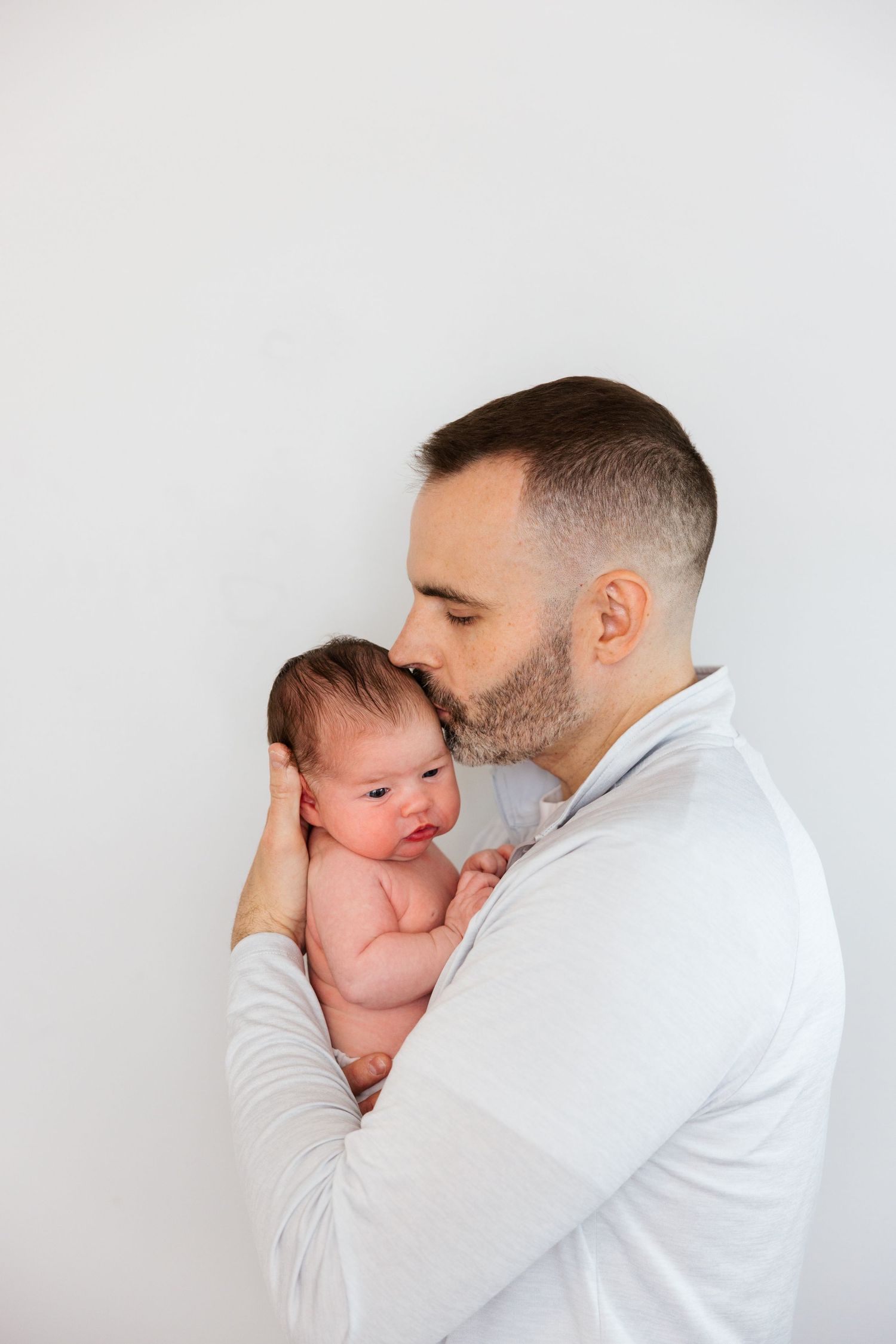 Father in white shirt embraces and kisses newborn baby against minimalist white background.