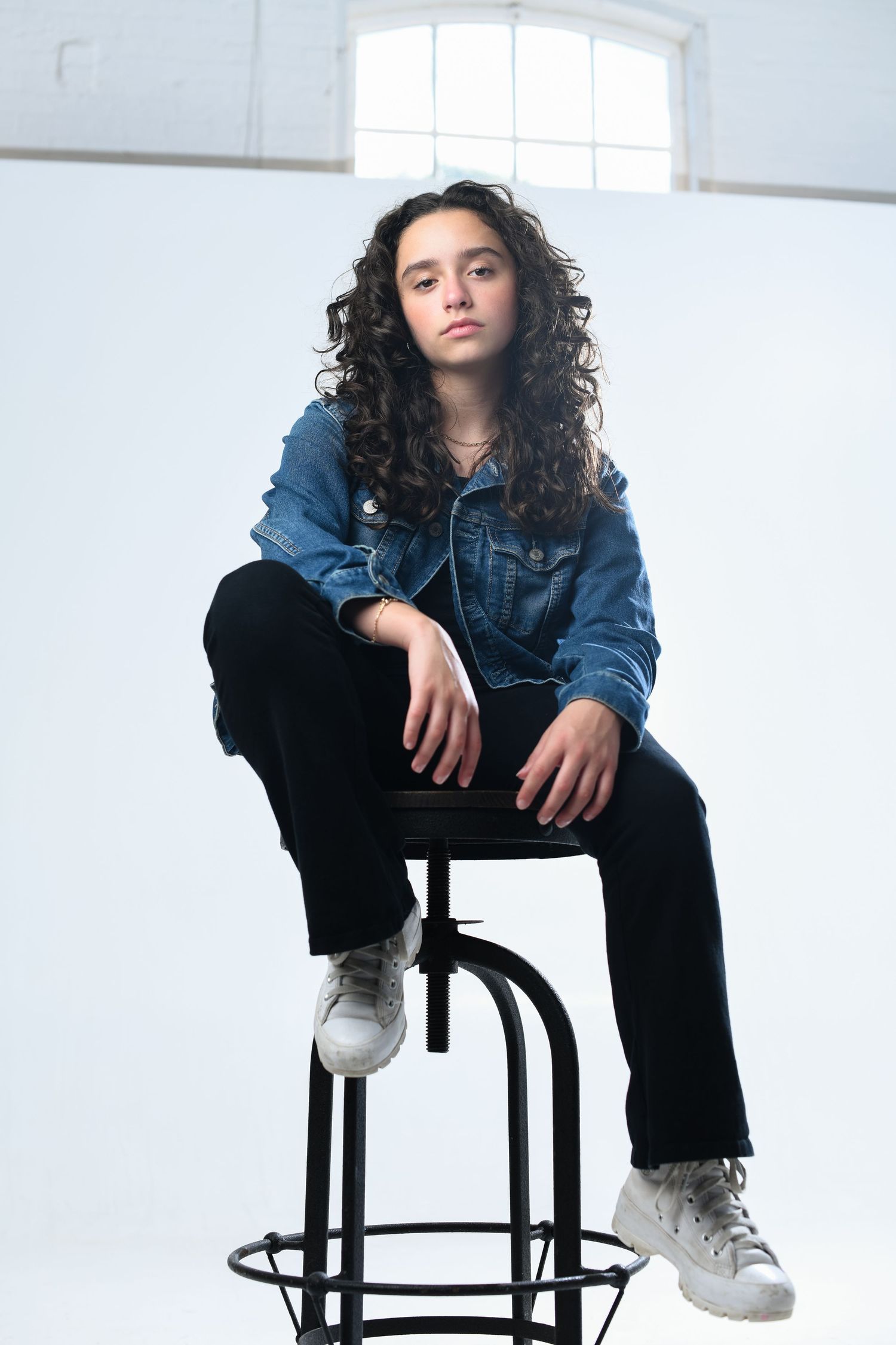 Young person with curly hair sitting on a stool, wearing a denim jacket, black pants, and white sneakers in a studio.