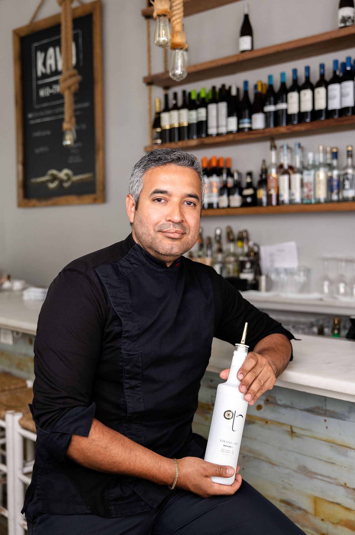 Man in black chef uniform holding a white bottle at a restaurant bar with wine bottles on shelves.