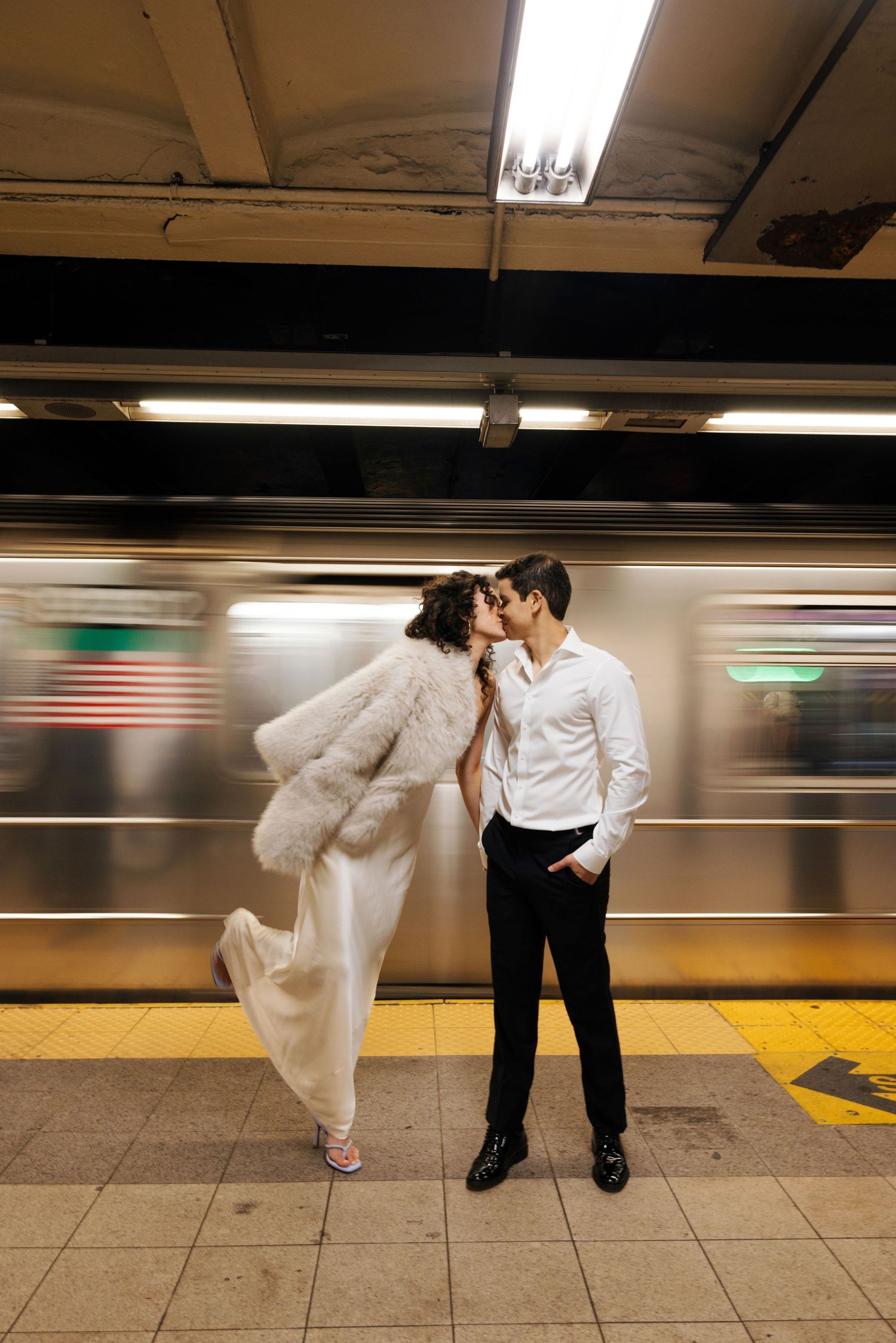 Couple sharing a romantic kiss on a NYC subway platform in wedding attire, with a blurred train passing behind them.