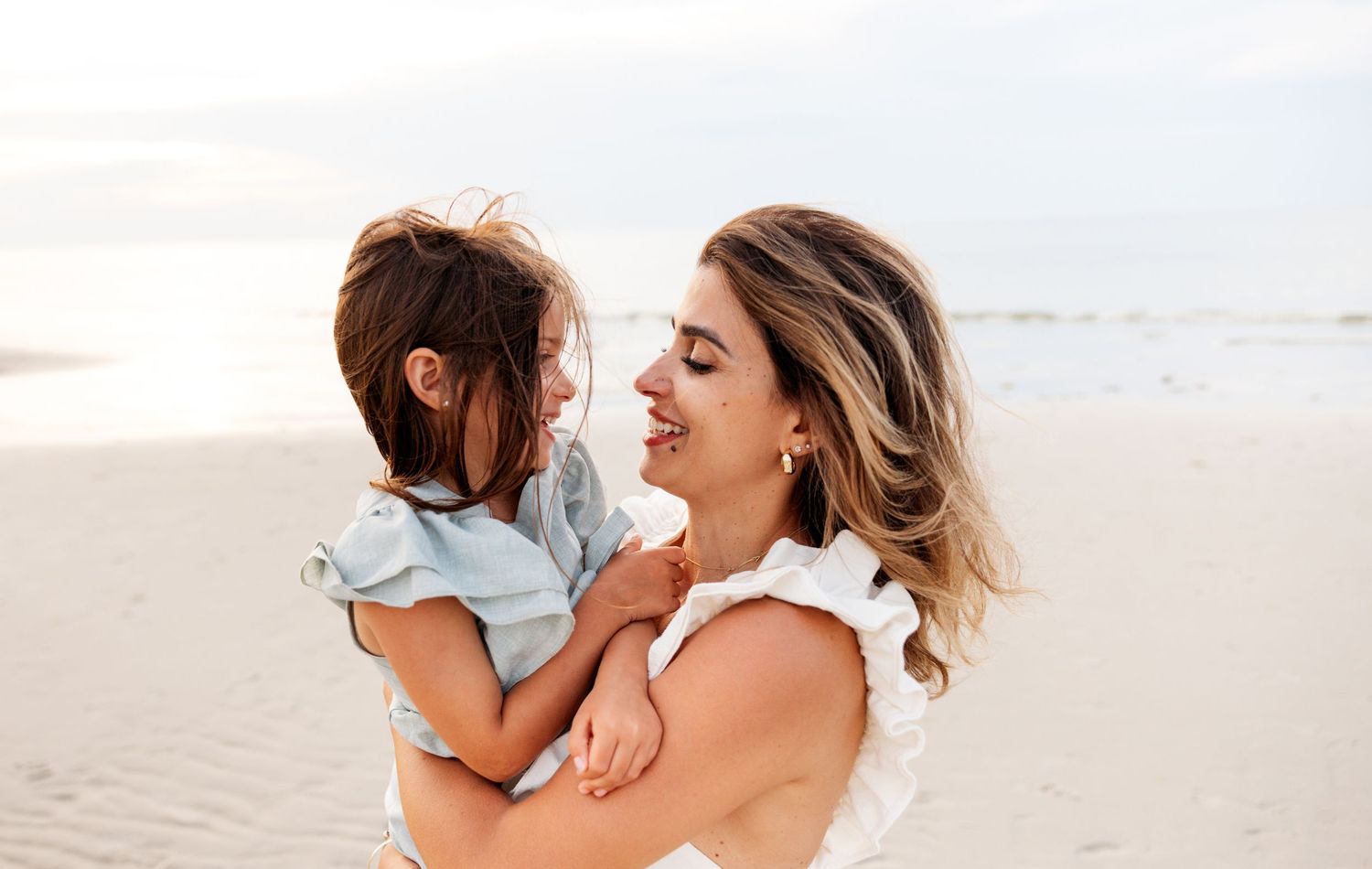 A beach goer in summer clothing holds a child while playing and laughing together on a white sandy shoreline.