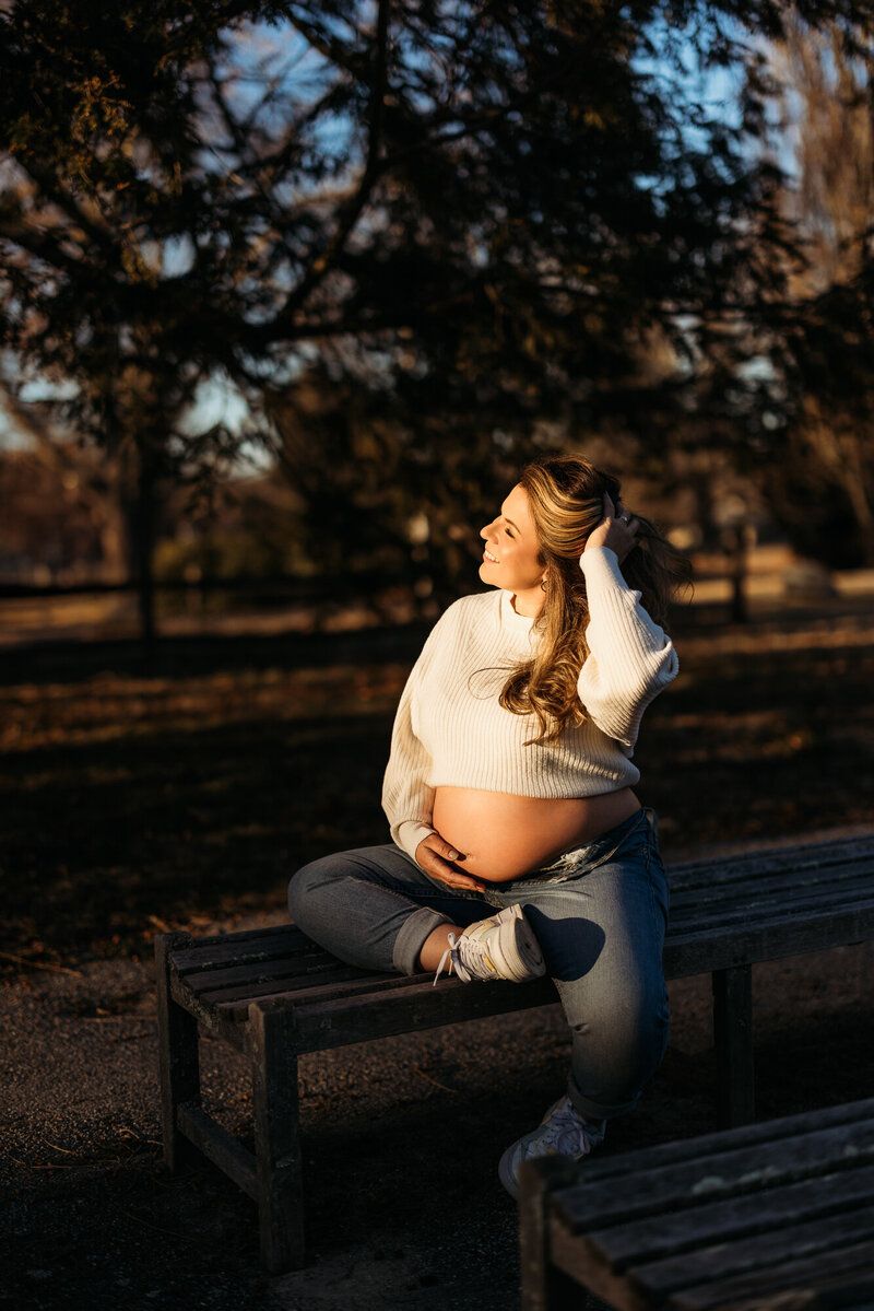 Woman in cream sweater sits on wooden bench during golden hour photoshoot.