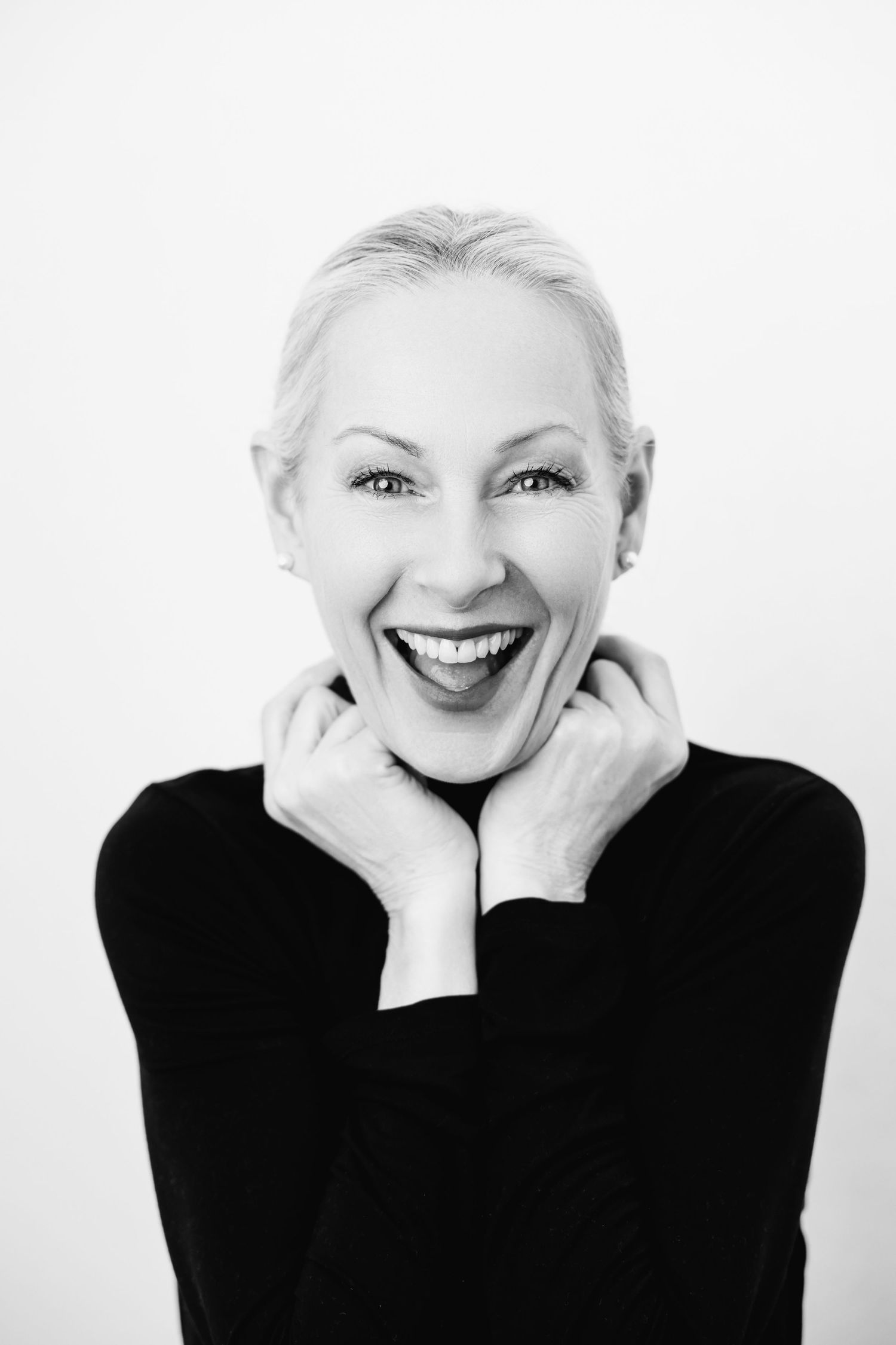 Black and white portrait of a joyful blonde woman in a black turtleneck, hands raised to chin, smiling broadly.