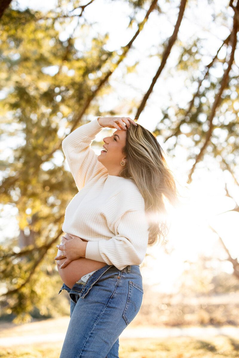 A woman in a cream sweater and jeans poses outdoors in golden autumn sunlight.