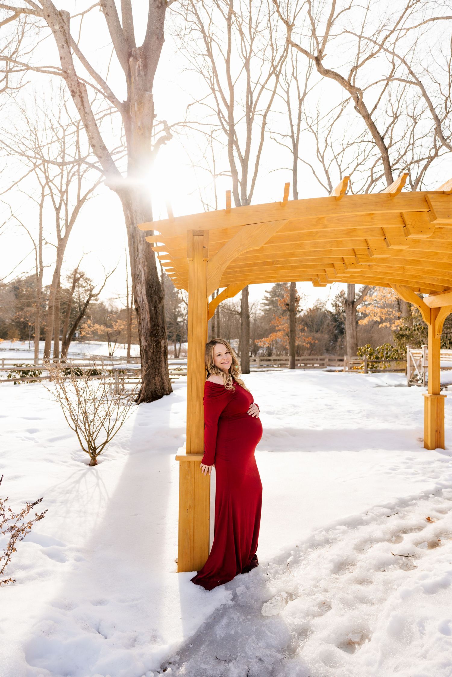 Winter maternity photos with woman in red dress by wooden pergola in snow.