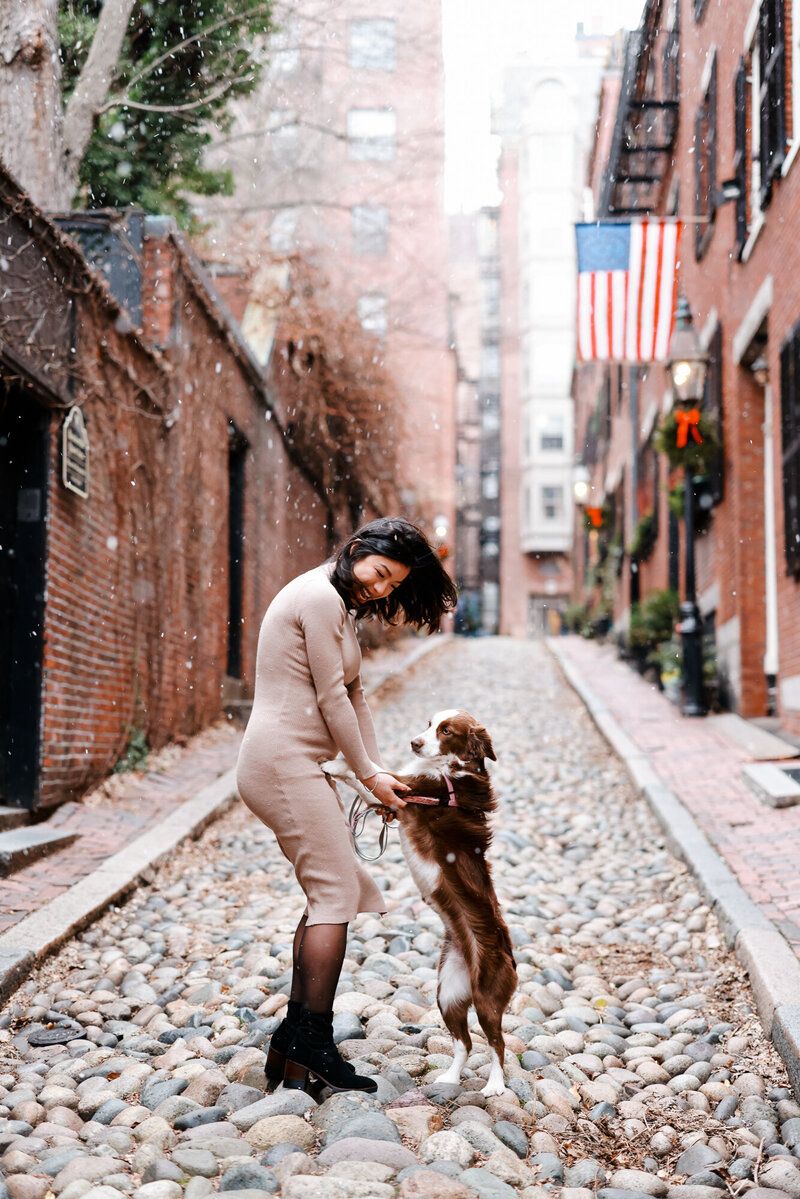 Person in beige dress playing with excited dog on historic cobblestone street with American flag visible.