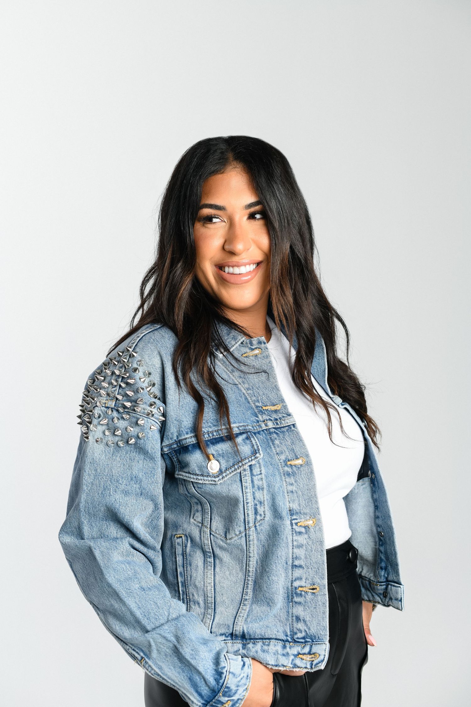 Smiling brunette woman in studded denim jacket and white top against gray background.