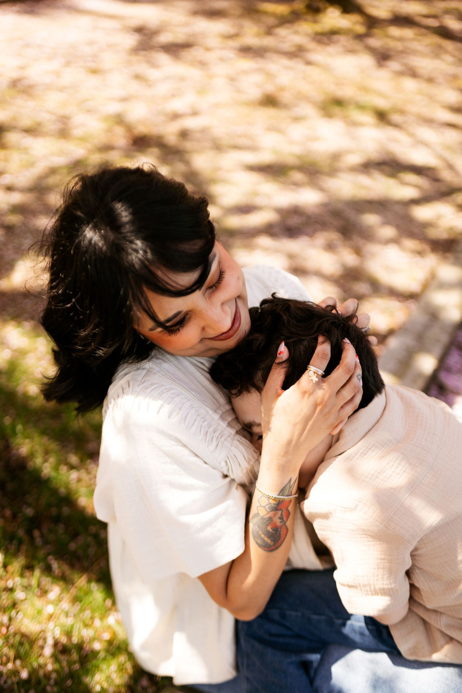 A tender moment shared between two people embracing outdoors in dappled sunlight, with one wearing white and having a tattoo.