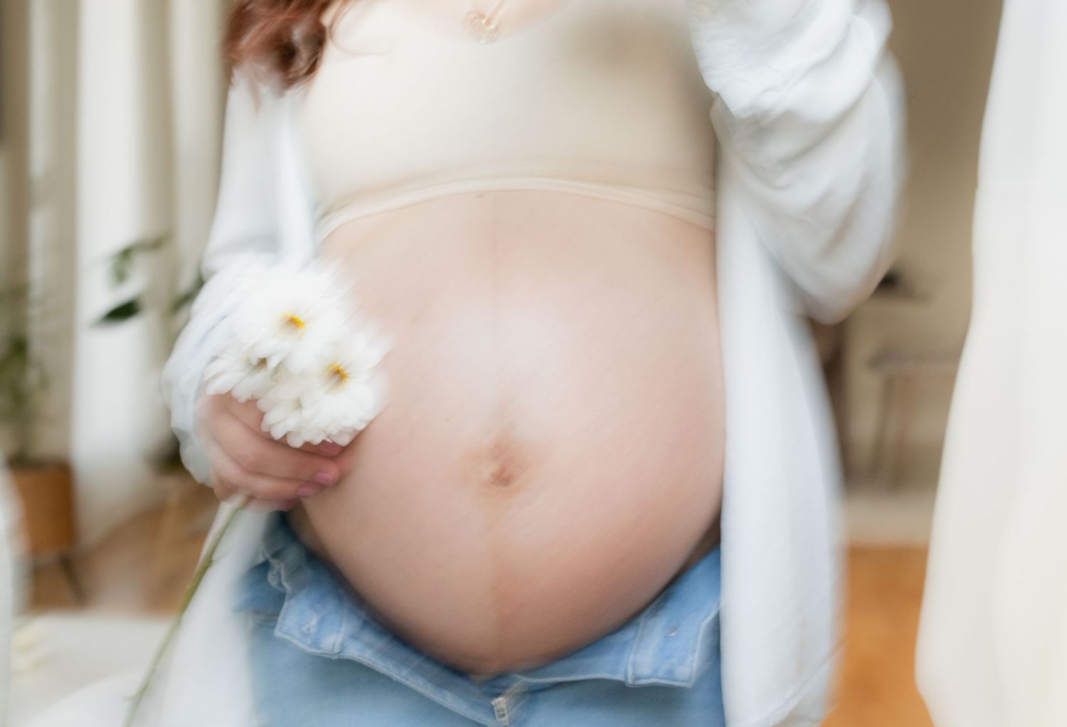 Pregnant woman in beige bralette and jeans holding white daisy flowers, showcasing baby bump in soft natural light.