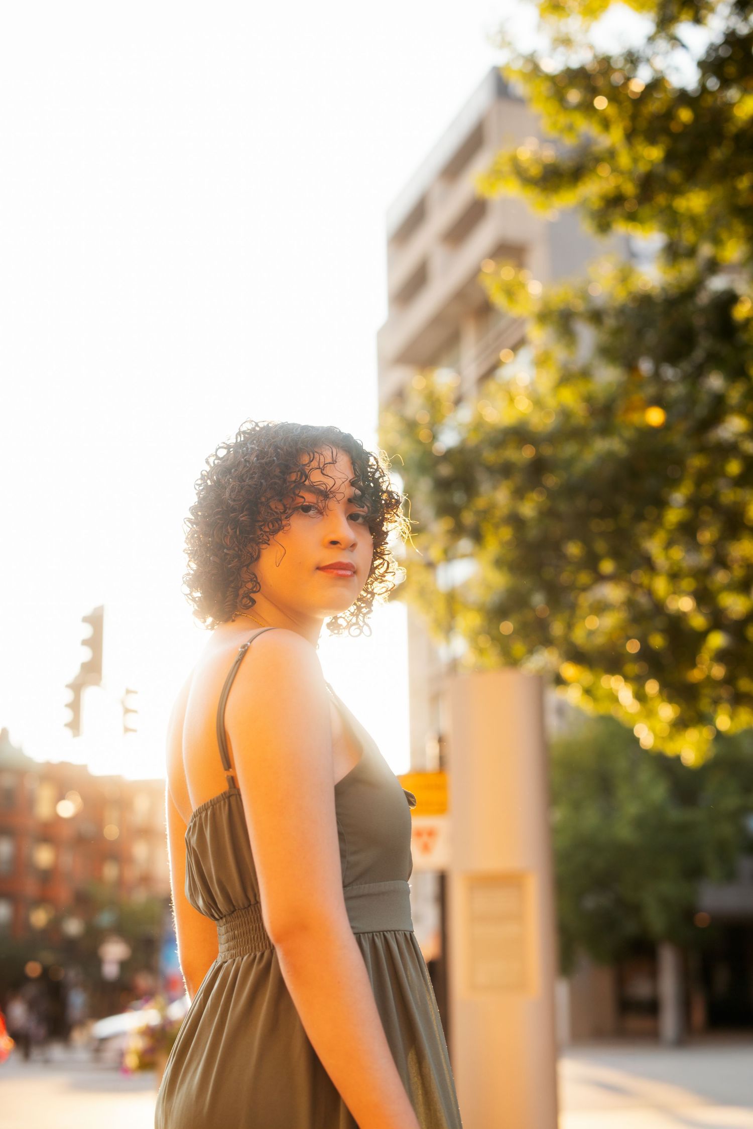 A person with curly hair poses outdoors during golden hour sunlight in an urban setting with trees and buildings visible.
