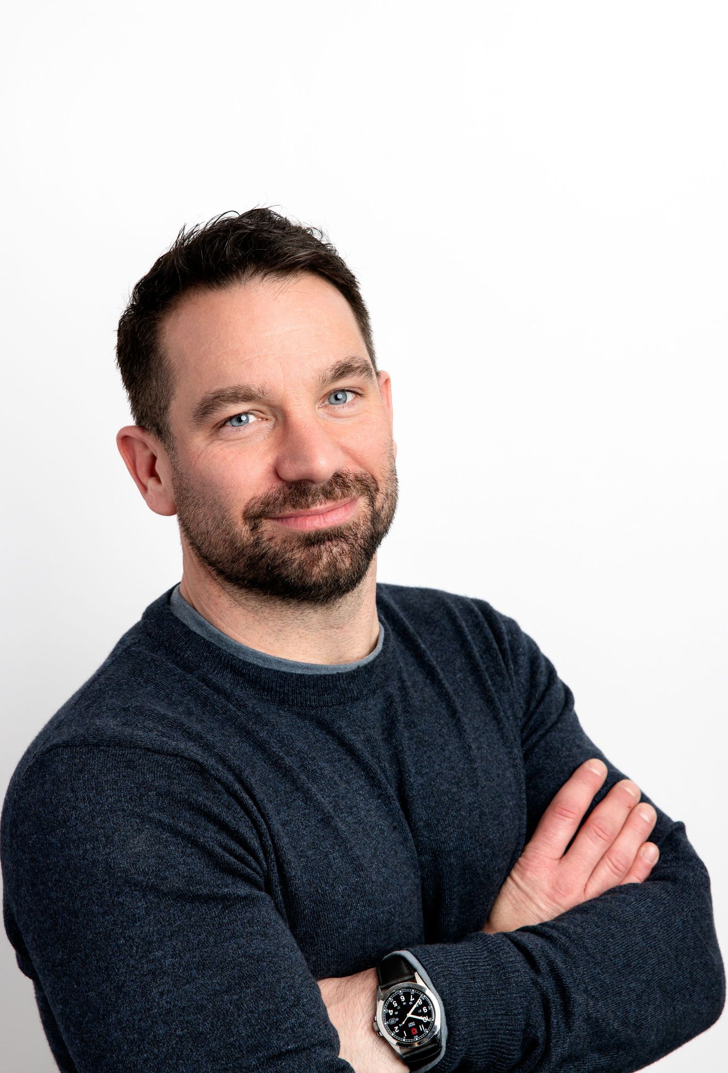 Smiling man with beard and dark hair, arms crossed, wearing navy sweater and watch, against white background.