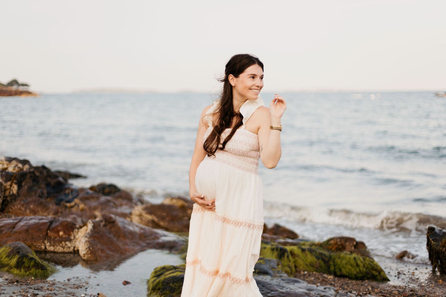 Woman in flowing peach dress walking along rocky coastal shoreline at sunset.